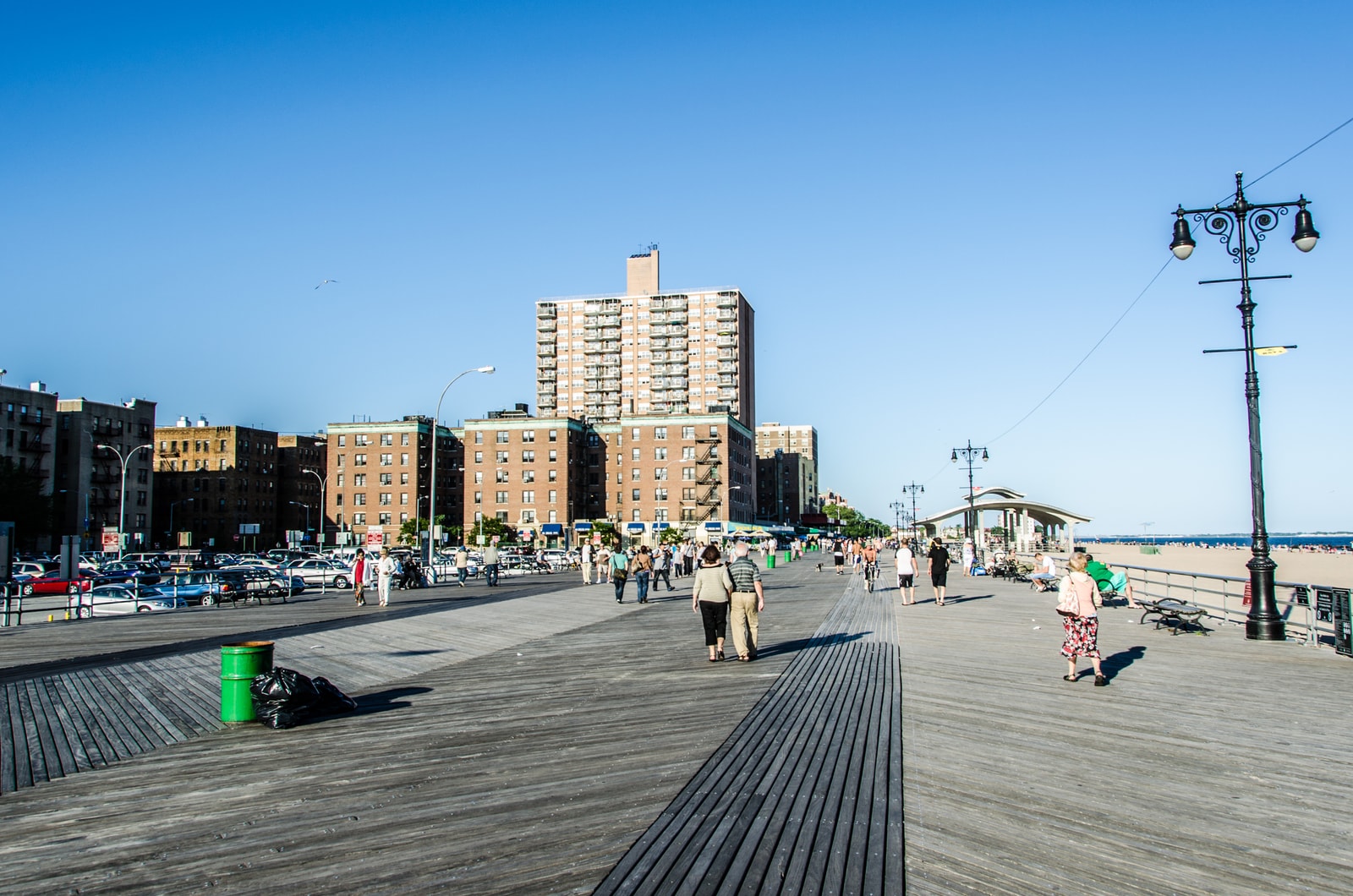 People strolling along the Riegelmann Boardwalk on a sunny summer day, with brick apartment buildings in the background.