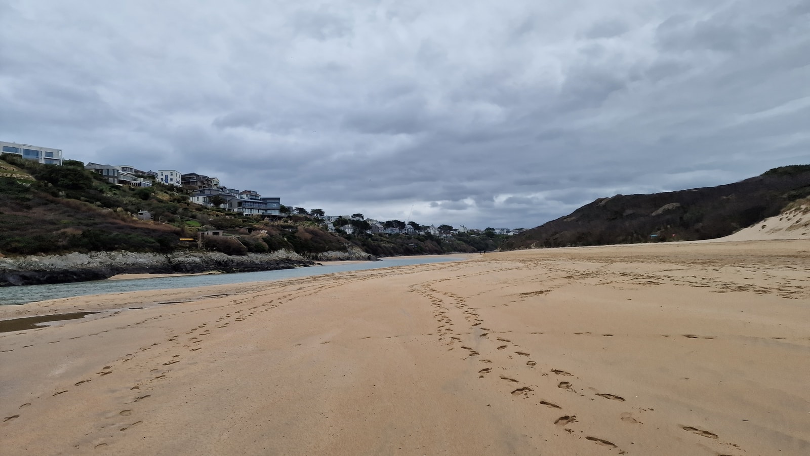 A wide view of the River Gannel estuary at low tide, with footprints in the sand and the river winding through.