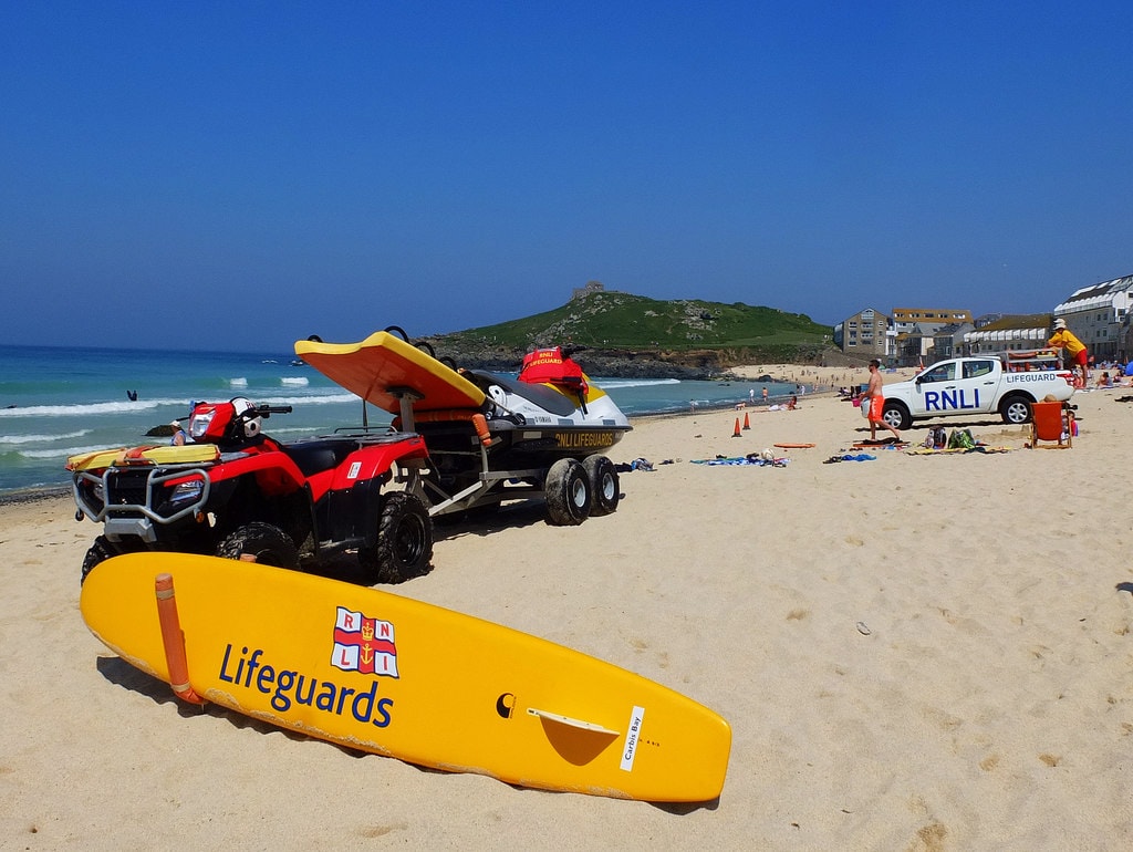 RNLI lifeguard equipment, including a red and yellow flag, rescue surfboard, and jet ski, is set up on the sand at Porthmeor Beach, ready for patrol.
