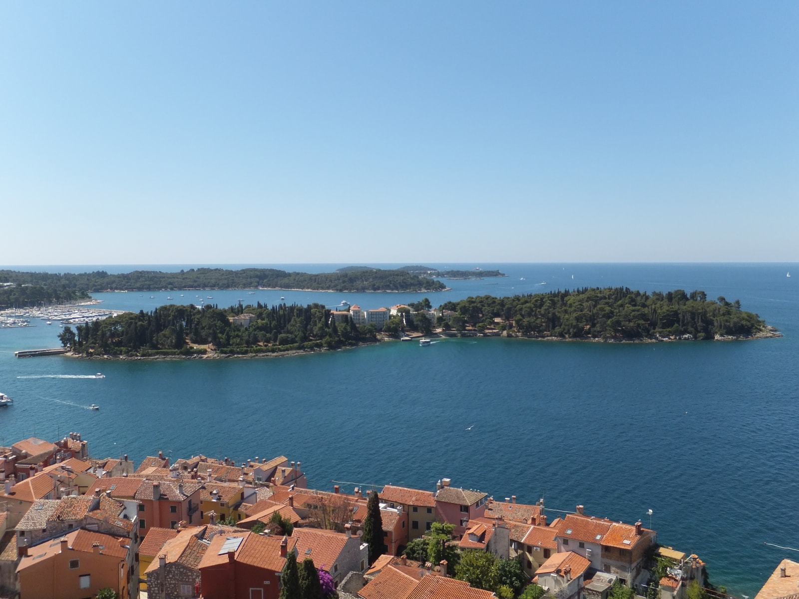 An aerial view from a bell tower looking down on the terracotta rooftops and historic center of Rovinj, with the marina and offshore islands visible in the distance.