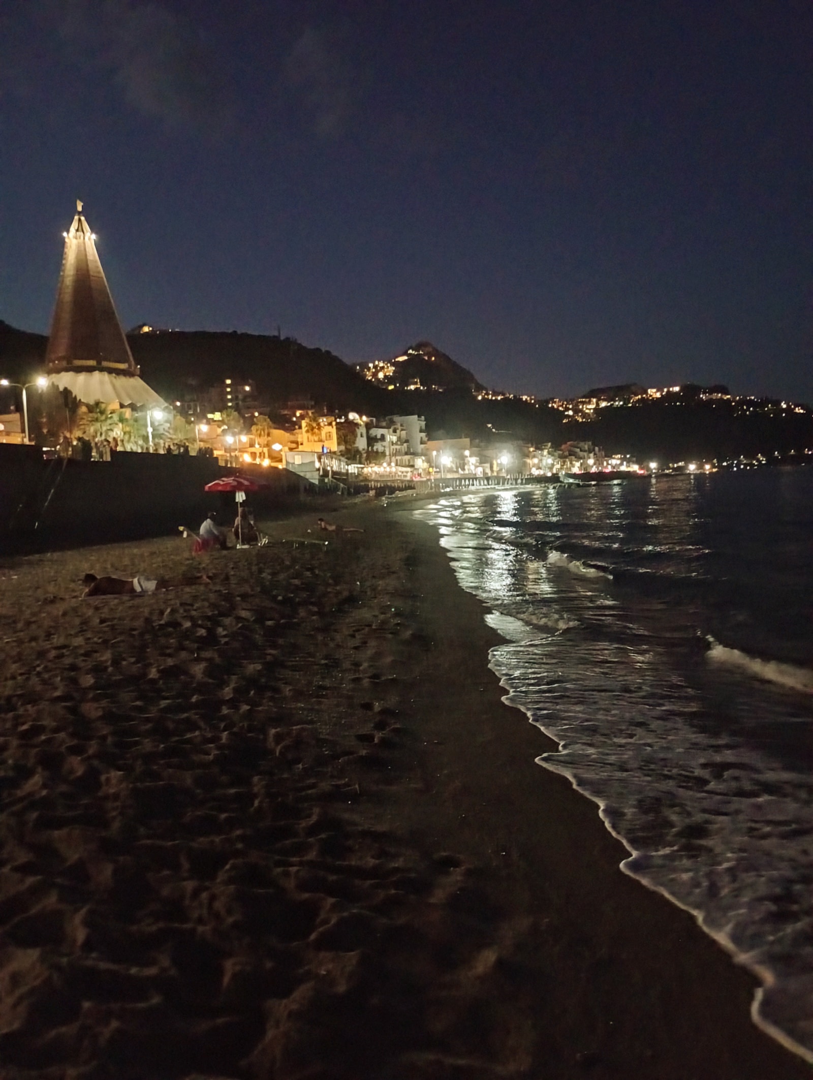 The warm lights of Giardini Naxos reflect on the sea at twilight, capturing the tranquil evening atmosphere of San Giovanni beach.