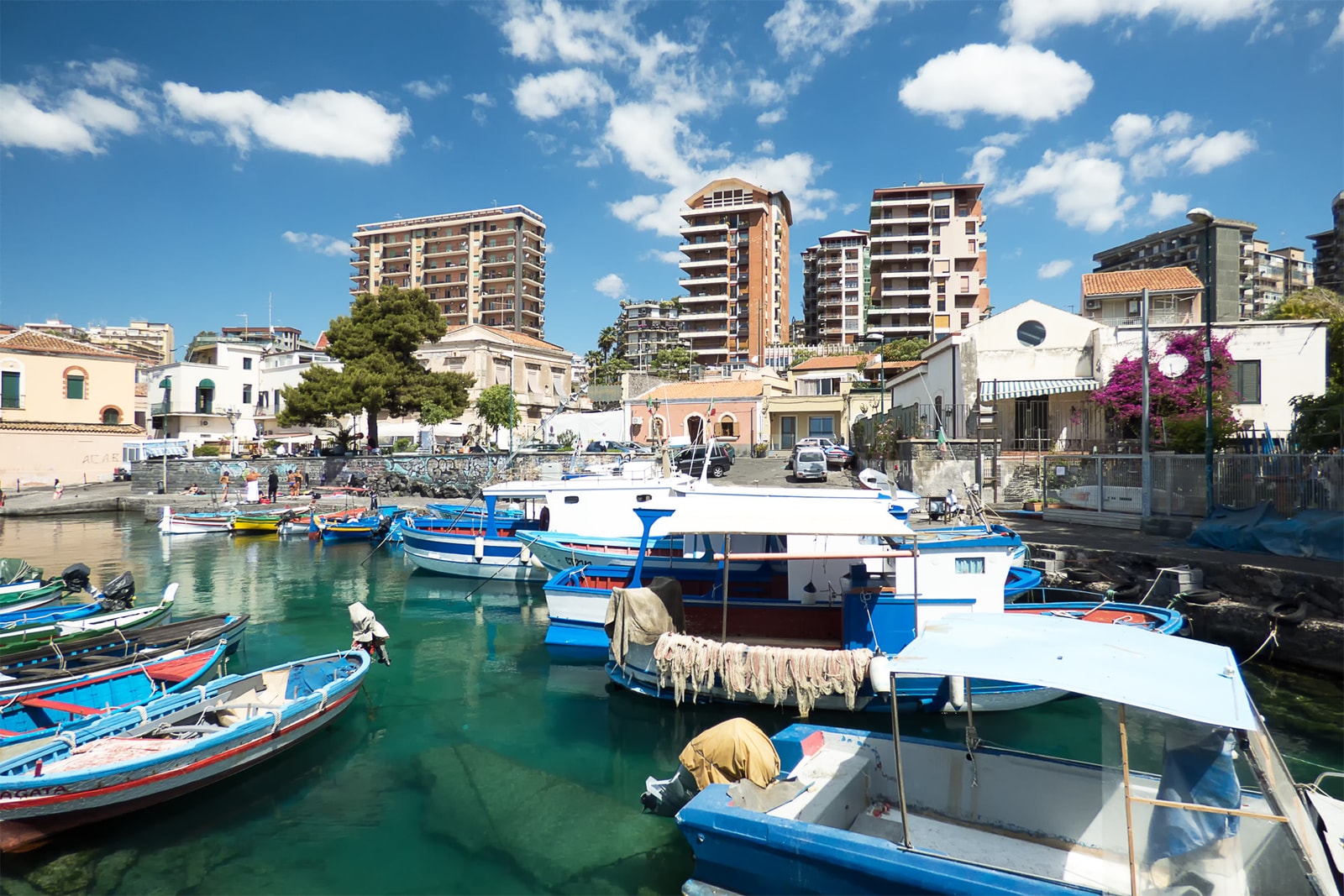 Small historic fishing harbor of San Giovanni Li Cuti with colorful wooden boats floating on clear water, contrasted with modern buildings behind.