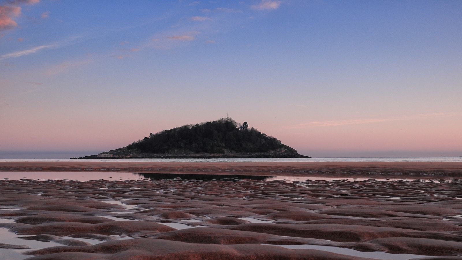 Santa Clara Island seen from La Concha Beach at sunset, with its silhouette framed by a pink and orange sky and ripples in the wet sand in the foreground.