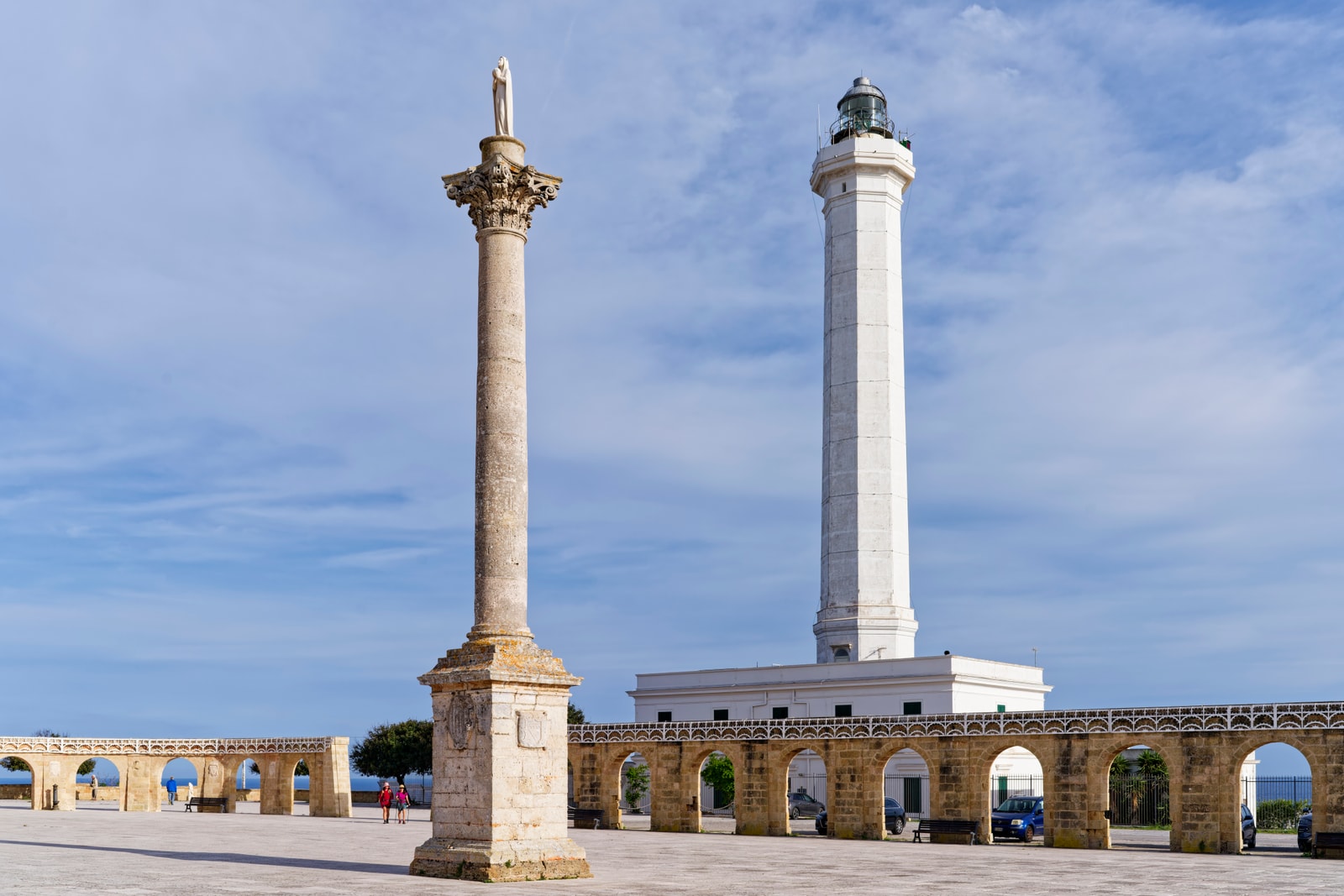 A view of the plaza in Santa Maria di Leuca, featuring its tall white octagonal lighthouse and a historic column against a blue sky.
