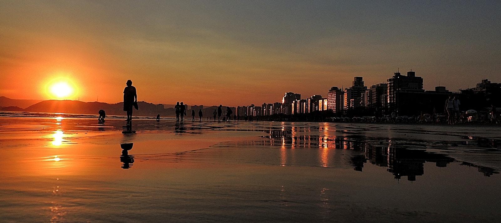 A wide panoramic view of the beachfront gardens in Santos at dusk, with the city skyline illuminated and reflecting in the wet sand.
