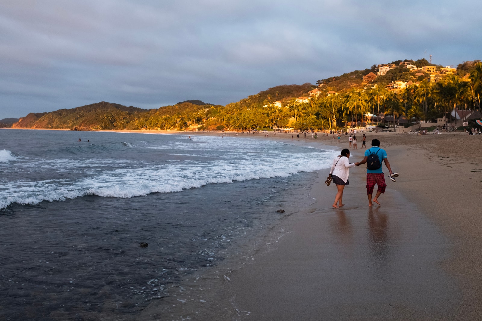 People walk along the wet sand of Sayulita beach as the waves recede, with lush green hills in the background.