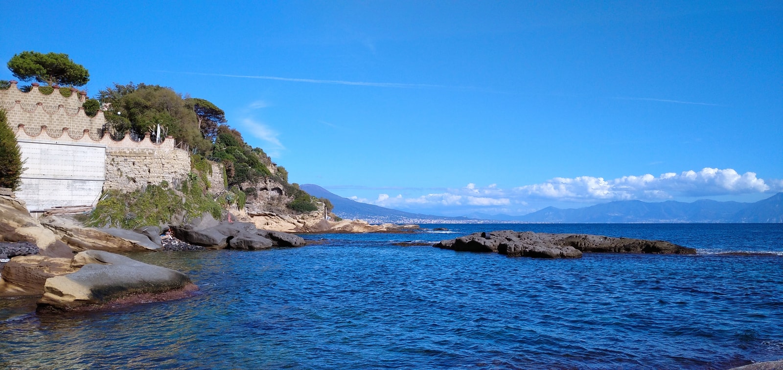 The volcanic rock platform of Scoglione at Marechiaro, with swimmers enjoying the water and the iconic silhouette of Mount Vesuvius visible across the bay.