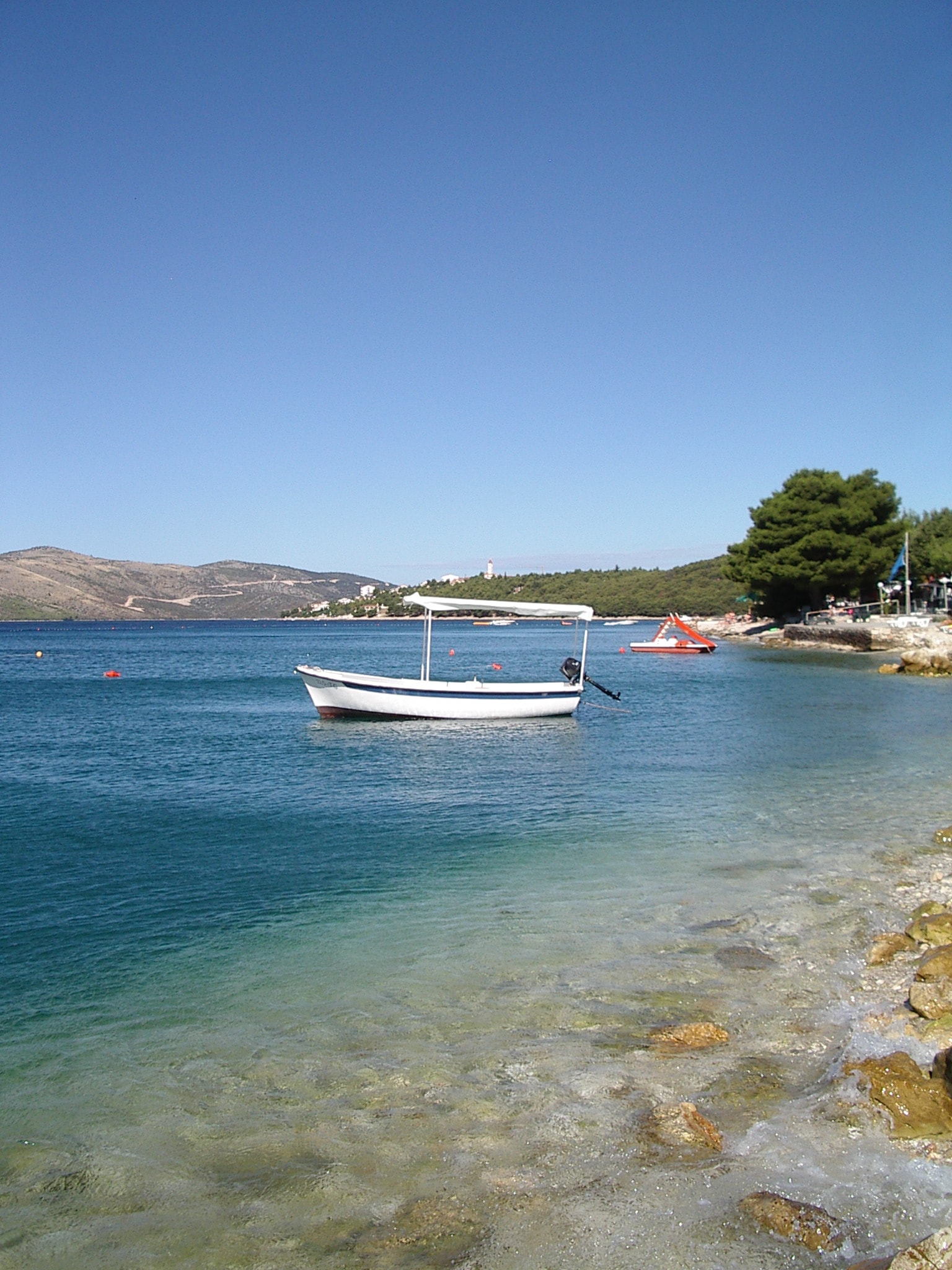 A small white boat rests in the clear, shallow turquoise water near a rocky Dalmatian shoreline.