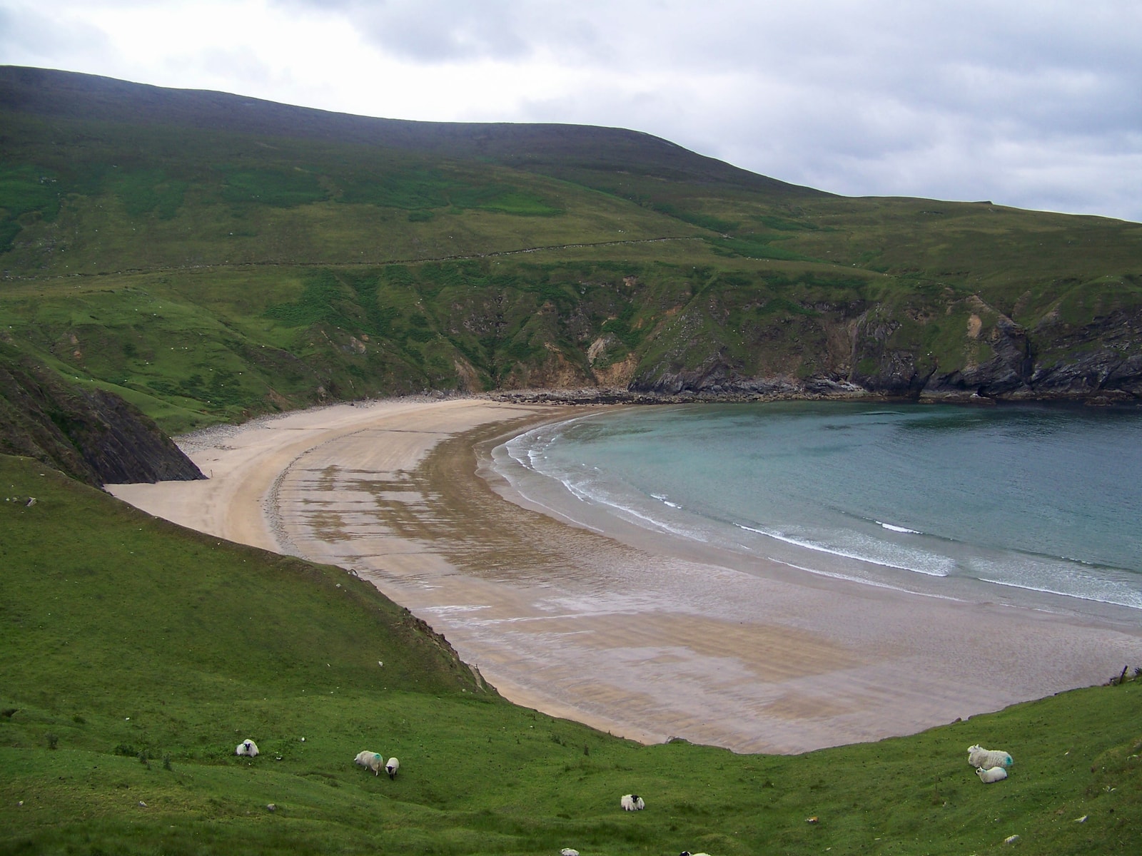 A high-angle view shows a perfect horseshoe-shaped beach with golden sand and emerald-blue water, nestled between steep green cliffs.