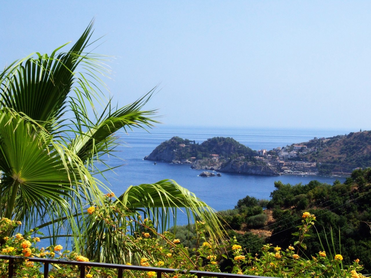 The serene Baia delle Sirene, or Siren's Bay, framed by tropical palm trees and colorful flowers against the bright blue Mediterranean Sea.