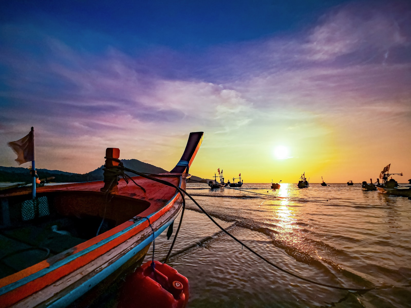 A longboat is silhouetted against a vibrant orange and purple sunset over the water in Sirinat National Park.