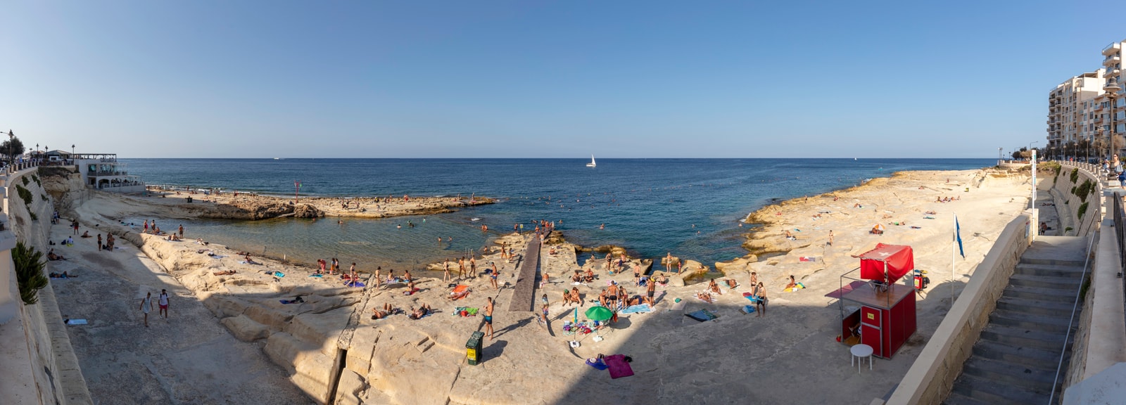 A panoramic view of the flat limestone platforms at Exiles Beach, packed with sunbathers and swimmers near the water's edge.