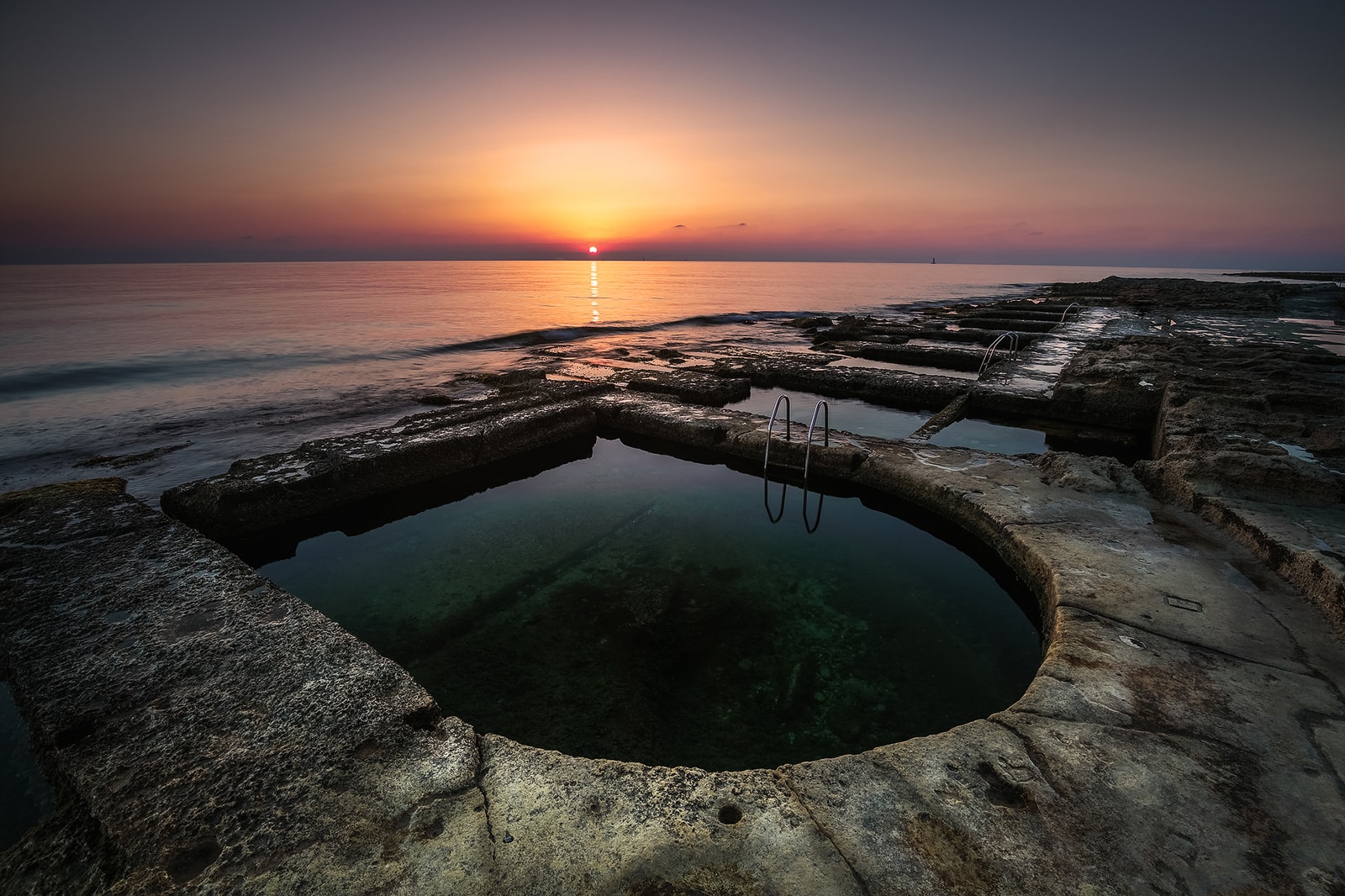 A close-up sunset shot of a round Victorian-era plunge pool carved into the rock at Sliema, with a metal ladder leading into the water.
