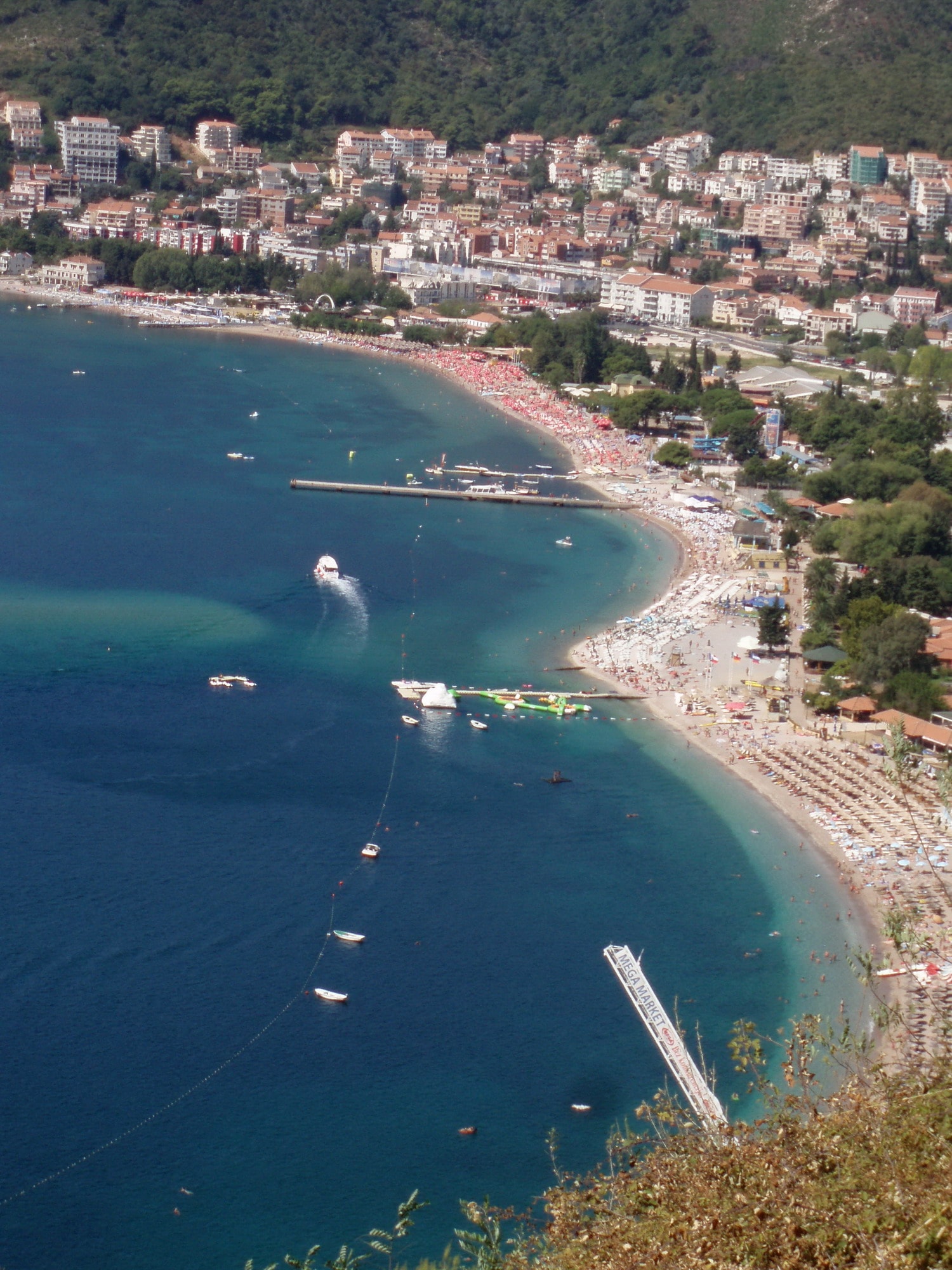 An aerial view shows a densely packed urban beach with rows of umbrellas, boats, and a seaside promenade.