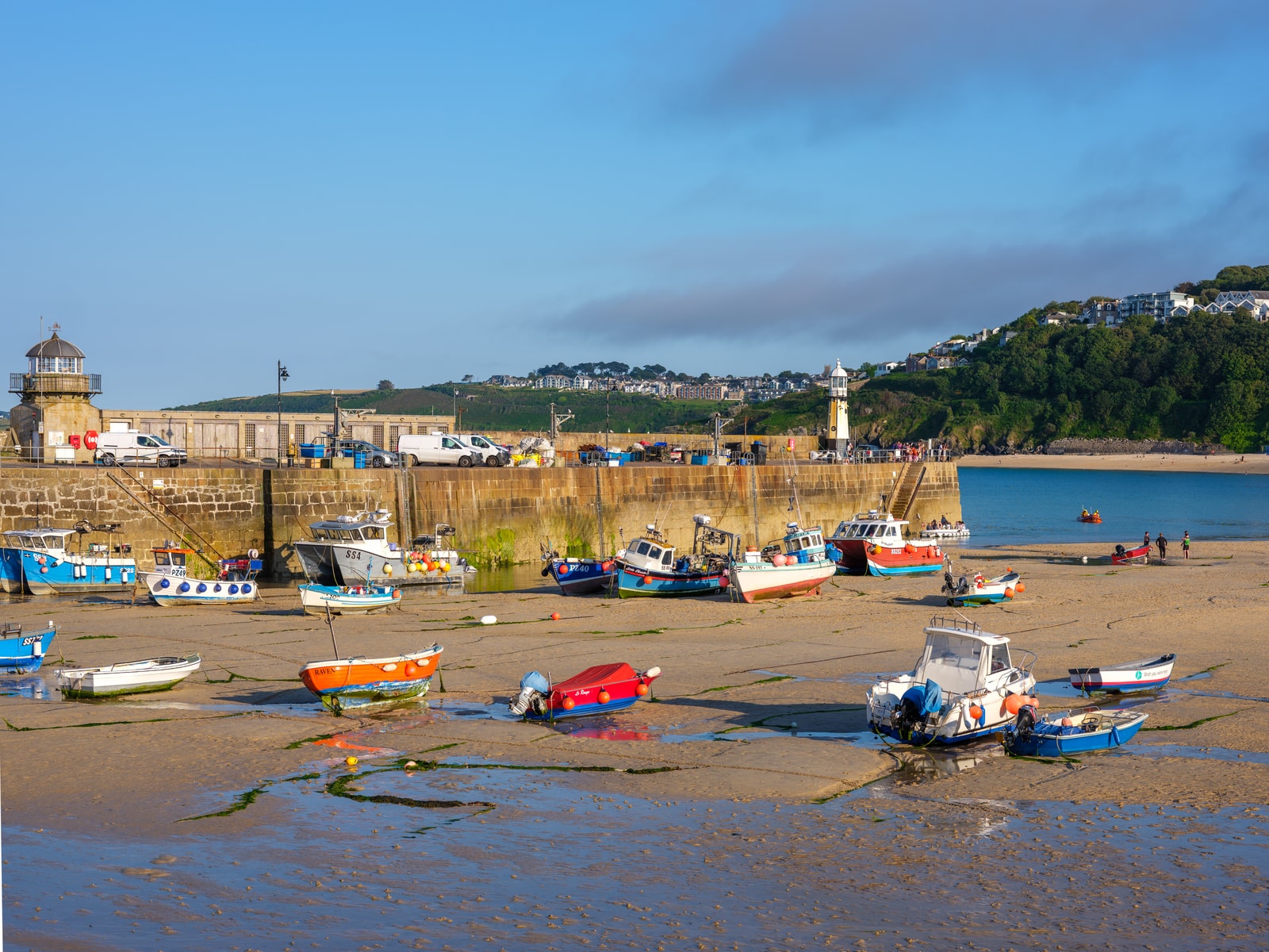Fishing boats are moored on the damp sand of the empty St Ives Harbour at low tide, with Smeaton's Pier and its lighthouse in the background.