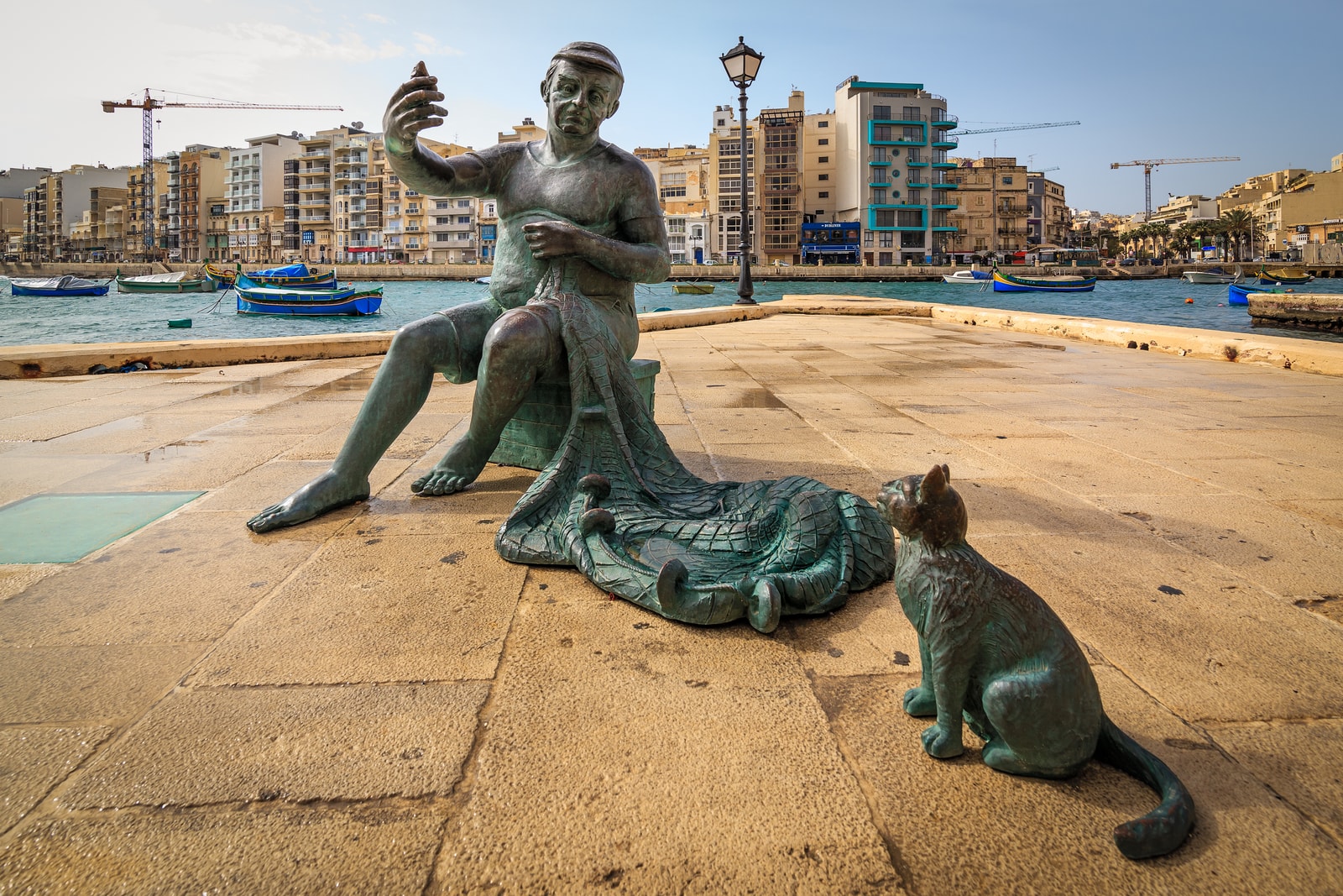A low-angle close-up of a bronze sculpture of a fisherman and a cat sitting on the stone quay in Spinola Bay, with traditional boats in the background.