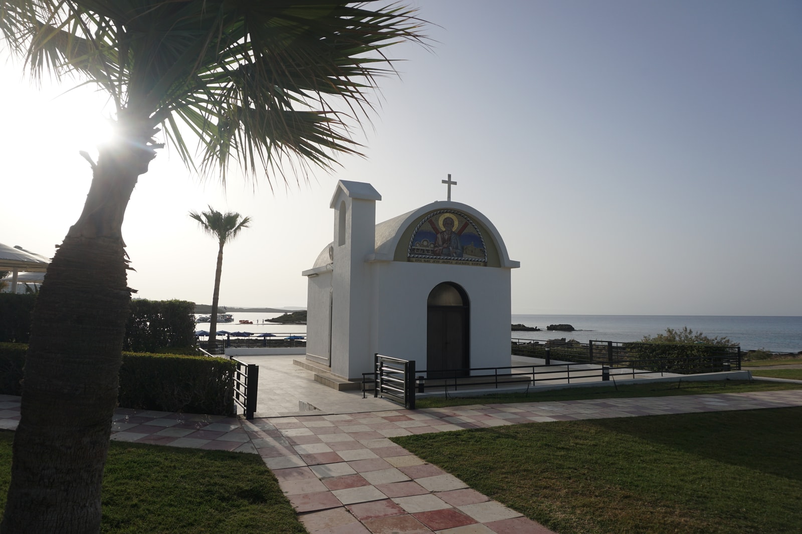 The traditional white St. Andrew's Church stands by Nissi Beach, framed by a palm tree and the blue sea.