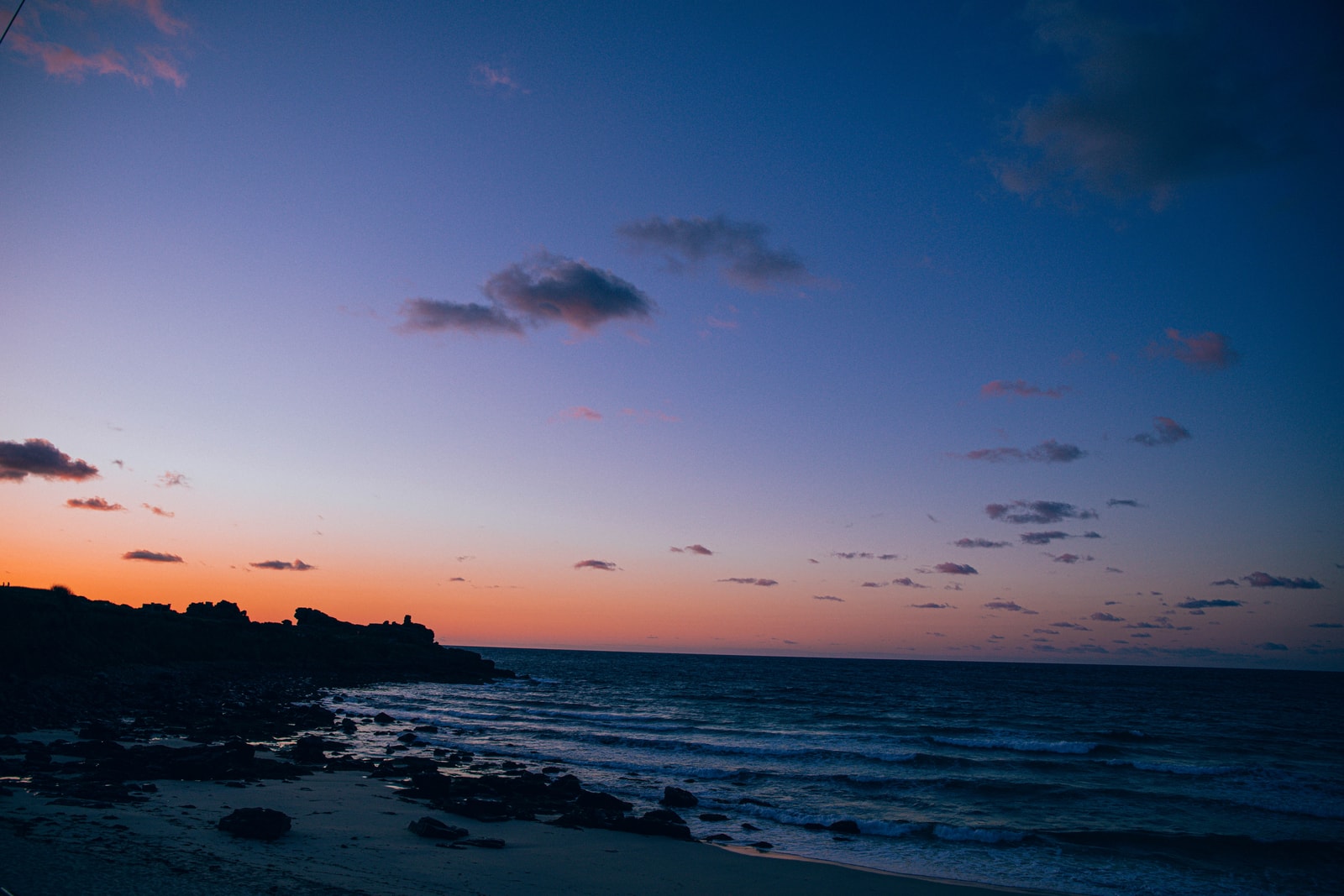 A vibrant sunset paints the sky with shades of orange and indigo over the silhouetted rocky coastline of St Ives Bay.
