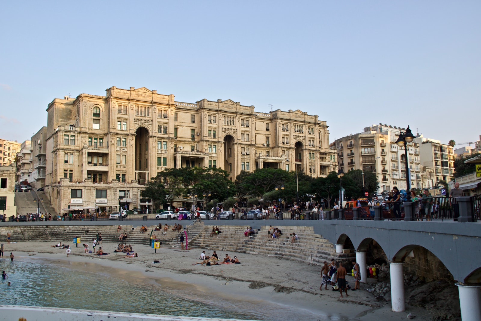 The small sandy beach at Balluta Bay, with people relaxing on the shore and the impressive Our Lady of Mount Carmel Parish Church in the background.