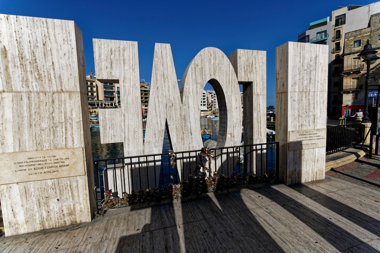 A close-up of the landmark LOVE monument on the Spinola Bay promenade, with couples' love locks attached to the fence in the foreground.