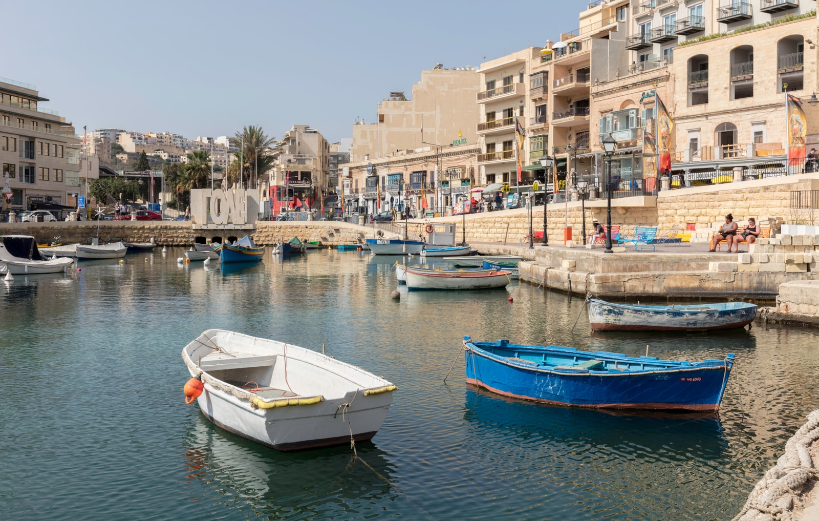 Vibrant, traditional Maltese luzzu fishing boats floating on the calm, turquoise waters of Spinola Bay, with seaside cafes in the background.