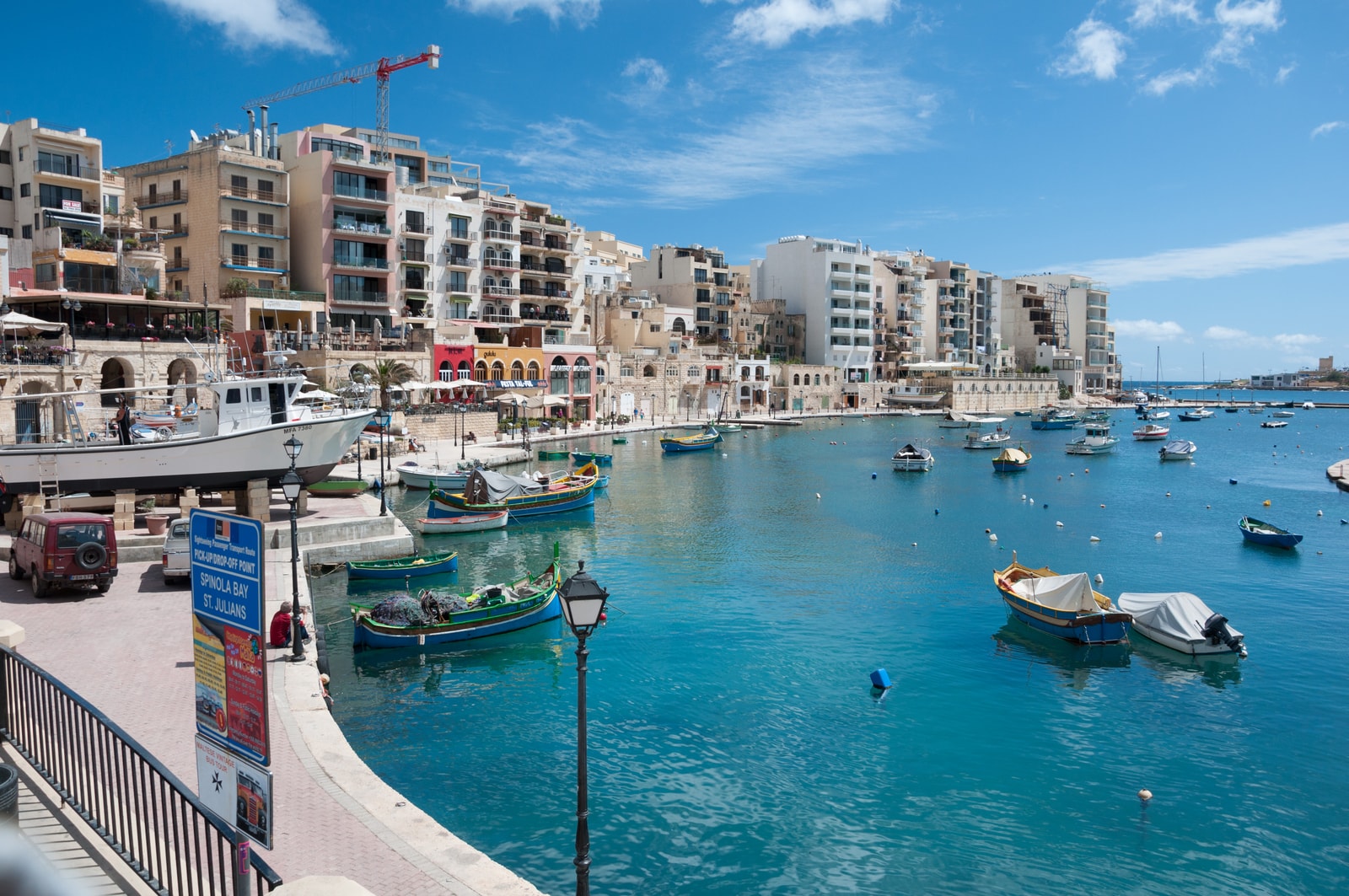 A crisp, sunny overview of Spinola Bay, showing traditional luzzu fishing boats anchored in the turquoise water with harborside apartments in the background.
