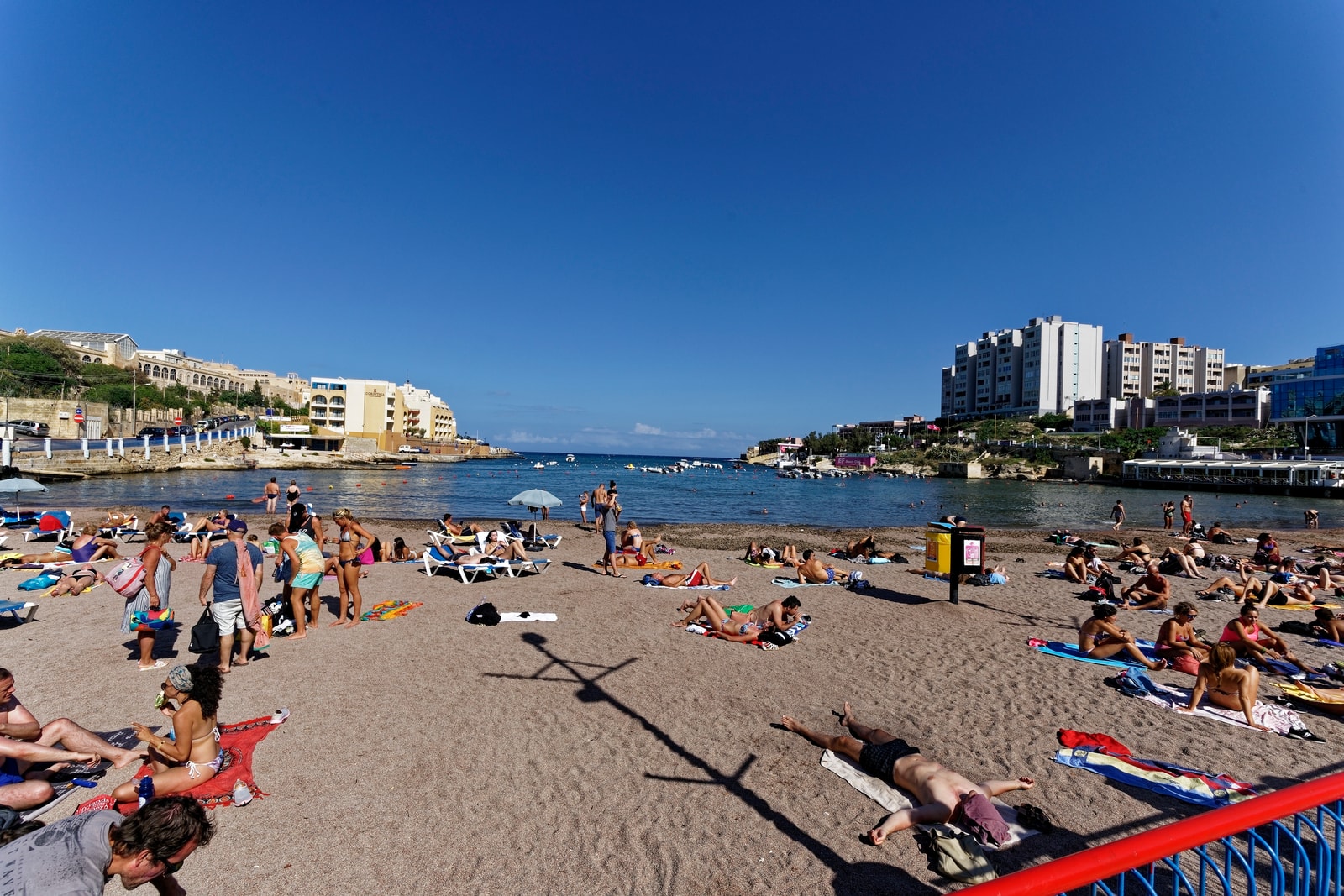 A sunny day at St. George's Bay, showing the sandy beach filled with sunbathers and colorful umbrellas, with hotels and resorts lining the calm blue water.