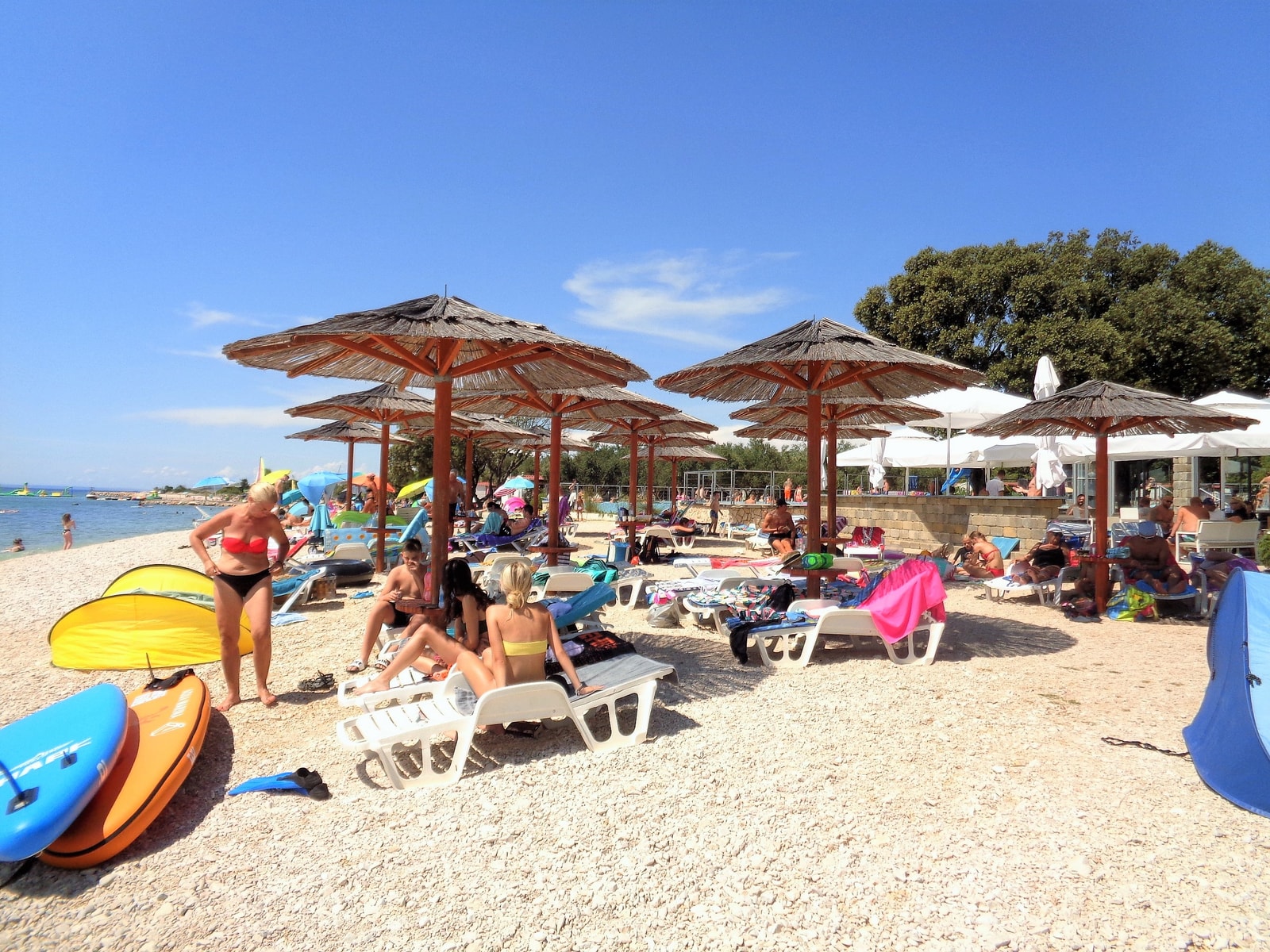 People relax on lounge chairs under thatched umbrellas on the pebbled shore of Straško Beach, with paddleboards nearby.
