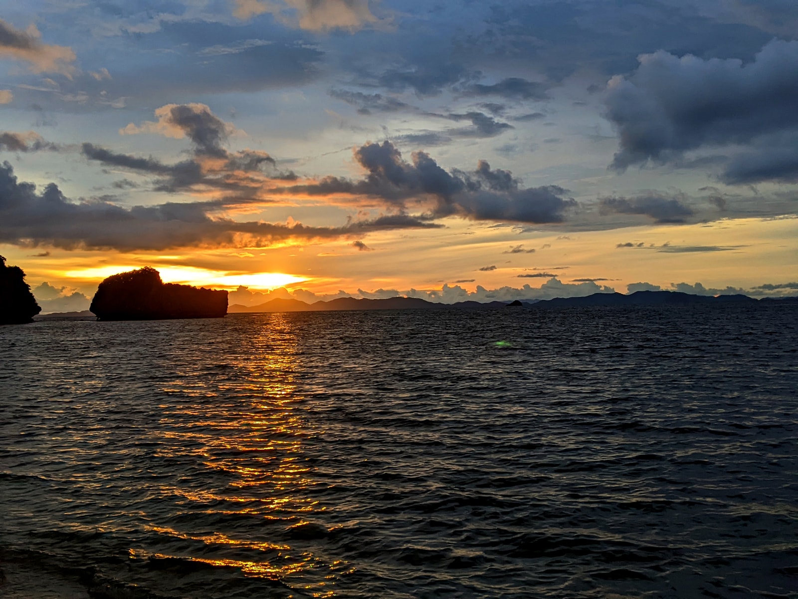 The setting sun casts a warm glow over the Andaman Sea, silhouetting distant islands and a longtail boat, as seen from a tranquil beach.