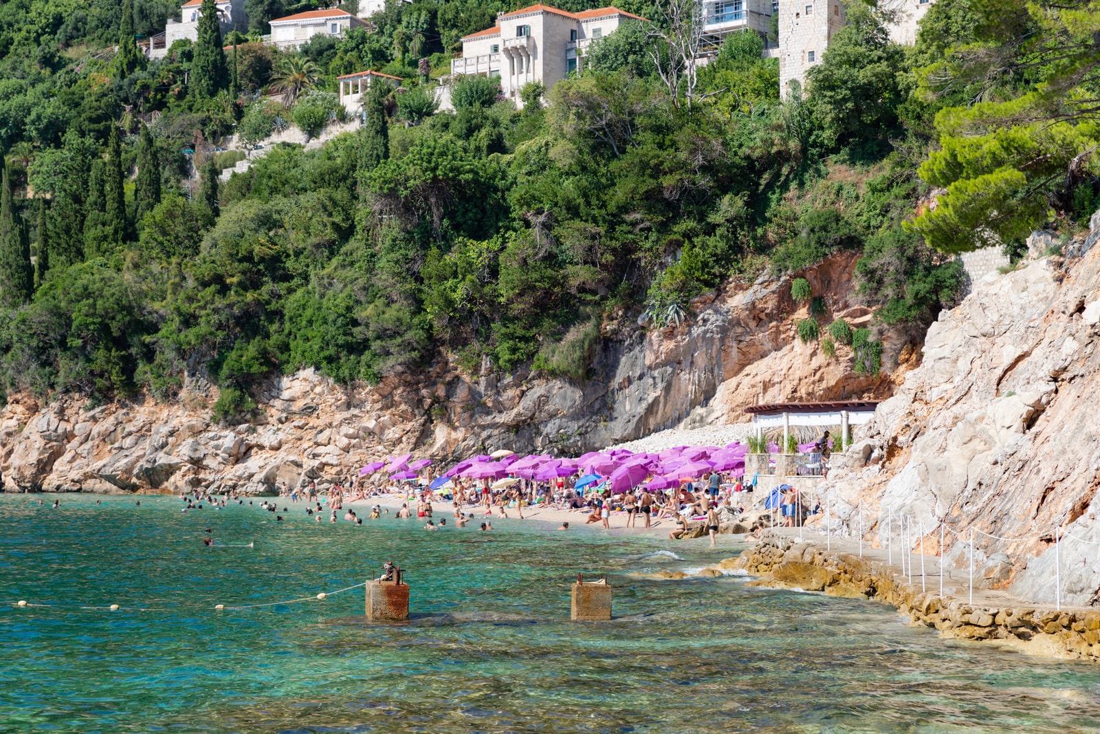 Sveti Jakov Beach is shown nestled between dramatic cliffs, with its signature purple beach umbrellas lining the shore.