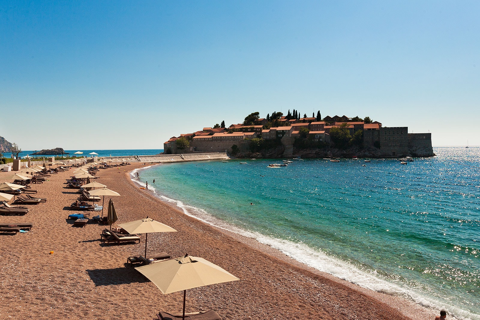 The shoreline in front of the island of Sveti Stefan, with loungers and umbrellas on a pebble beach.
