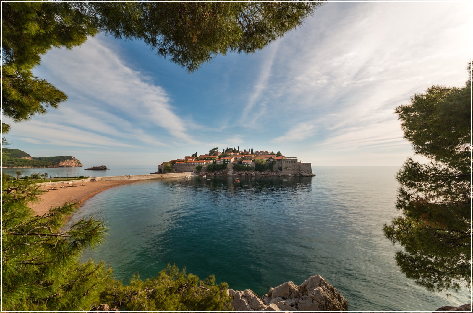 A scenic view frames the island of Sveti Stefan across a turquoise bay, with green foliage in the foreground.