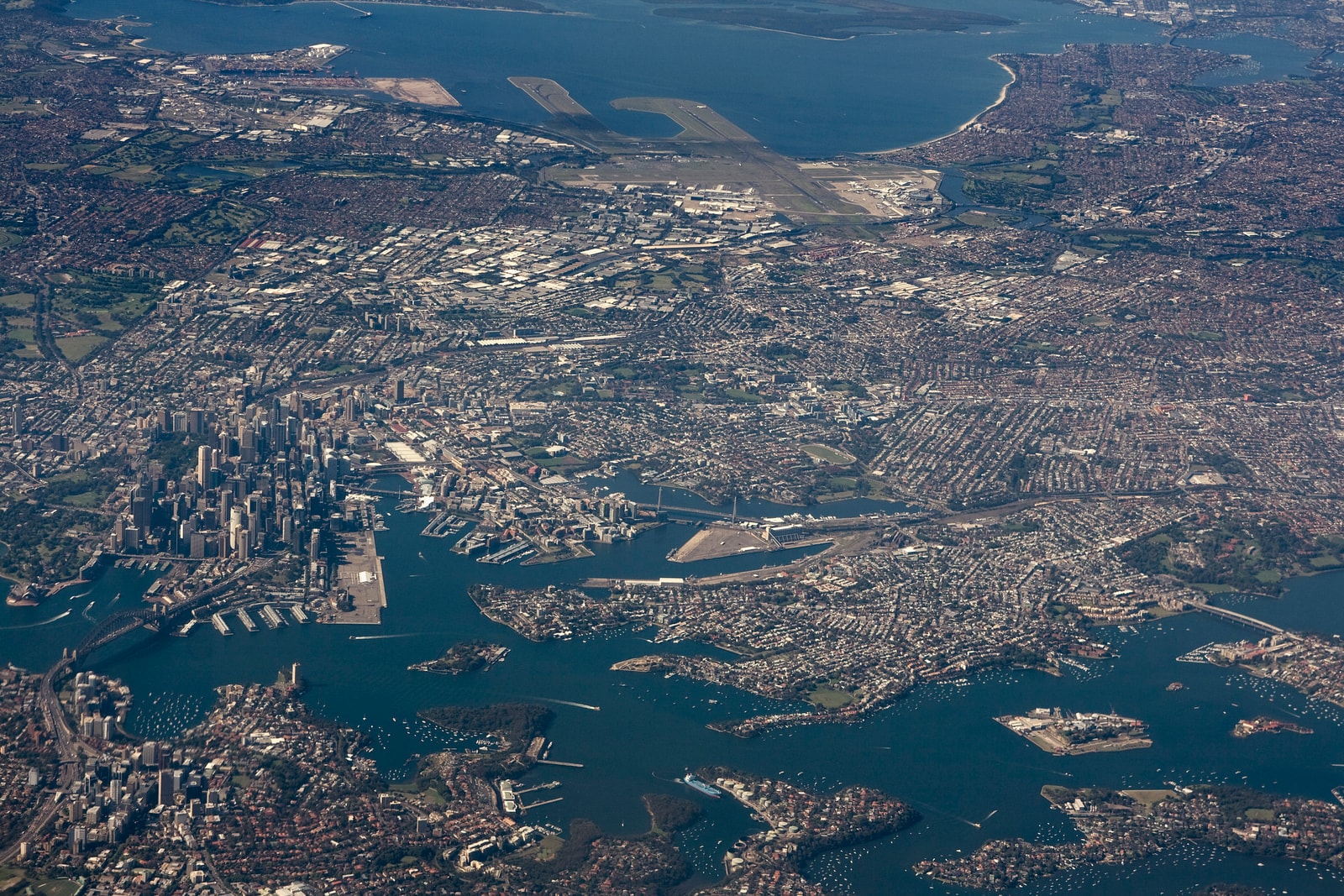 A broad aerial photograph showing the complex geography of Sydney, with its harbour, bays, coastline, and dense suburban areas.
