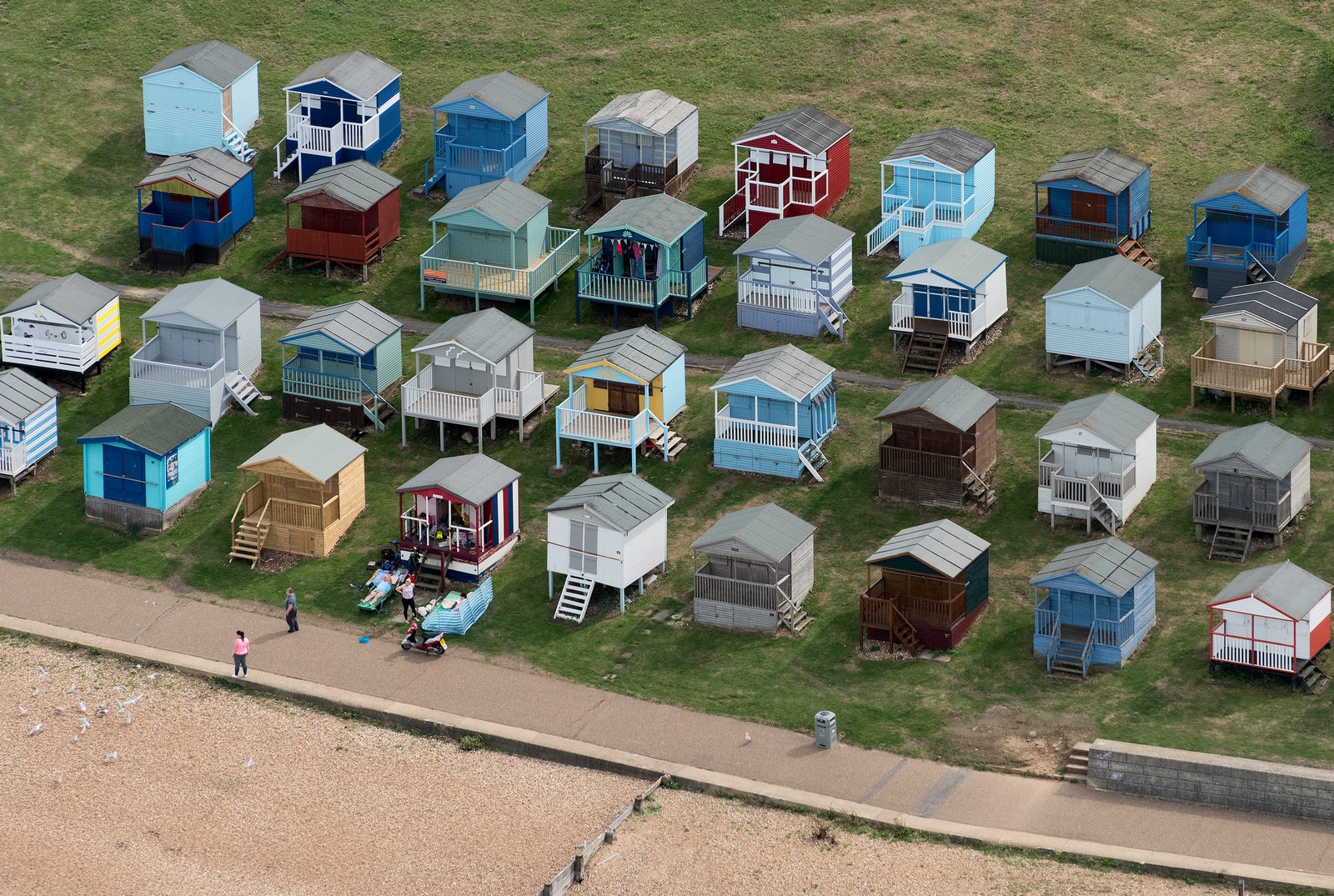 An aerial view of colorful pastel beach huts arranged on a grassy slope next to a pebble beach.