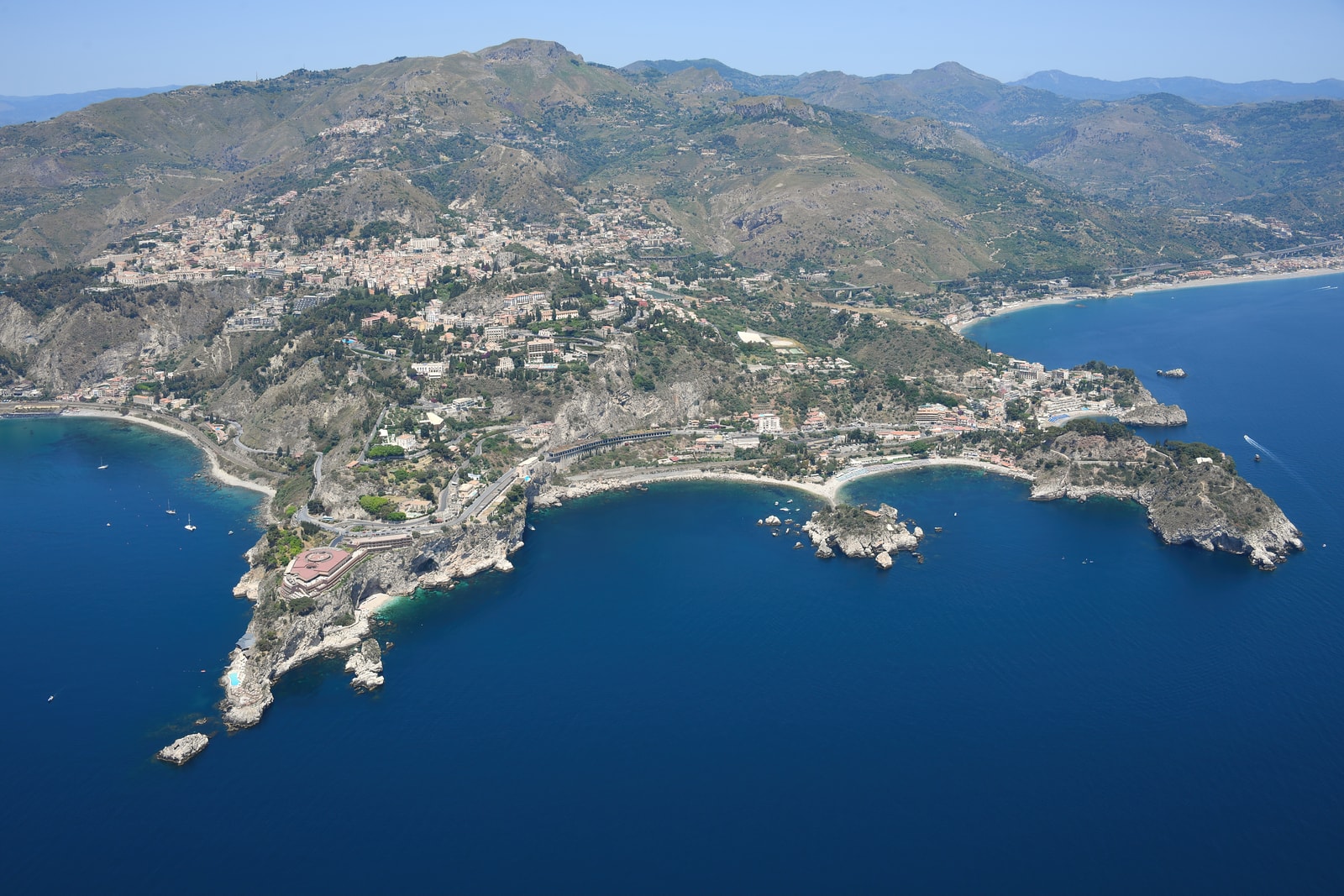 High altitude aerial view of Taormina's dramatic cliffs, winding coastal roads, and deep-blue bays from the southeast.