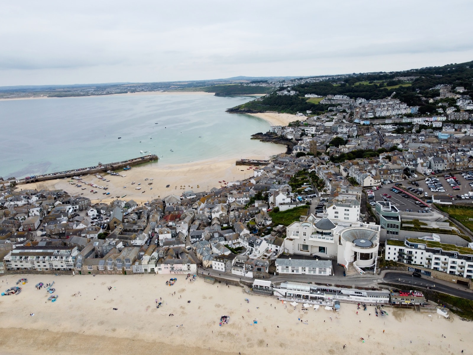 An aerial view shows the iconic circular building of the Tate St Ives gallery situated directly behind the golden sands of Porthmeor Beach, with waves rolling in from the Atlantic.