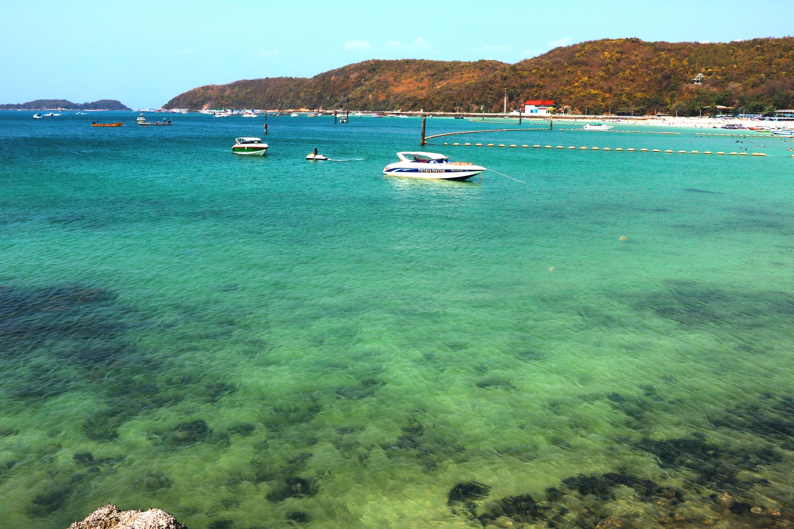 Boats anchor in the clear, turquoise water of Tawaen Beach on Koh Larn, with a white sandy shore and green hills behind.