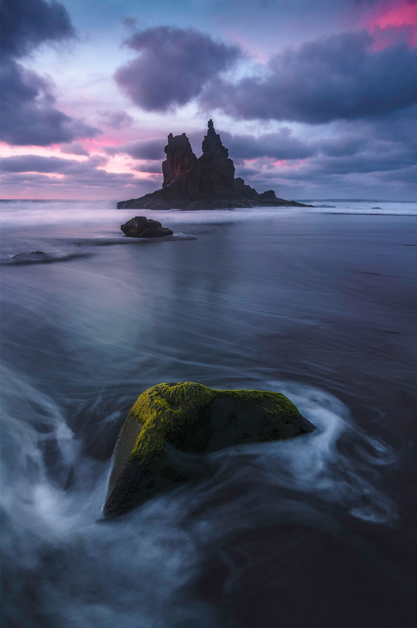 The dark sand of Playa de Benijo glows during a colorful sunset, with dramatic sea stacks silhouetted against the water.