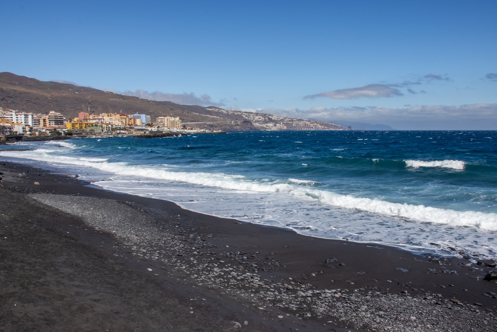 A view of a black sand beach with breaking waves, with a coastal town built on the hills in the background.