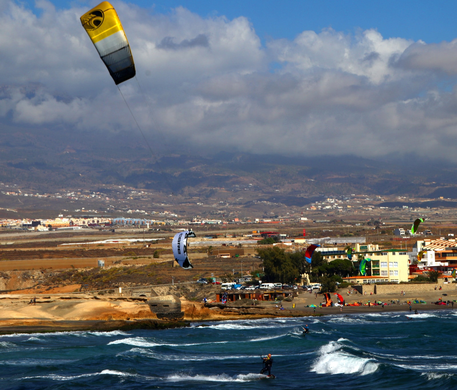 Several kitesurfers are on the choppy water of El Médano, with their colorful kites in the air.