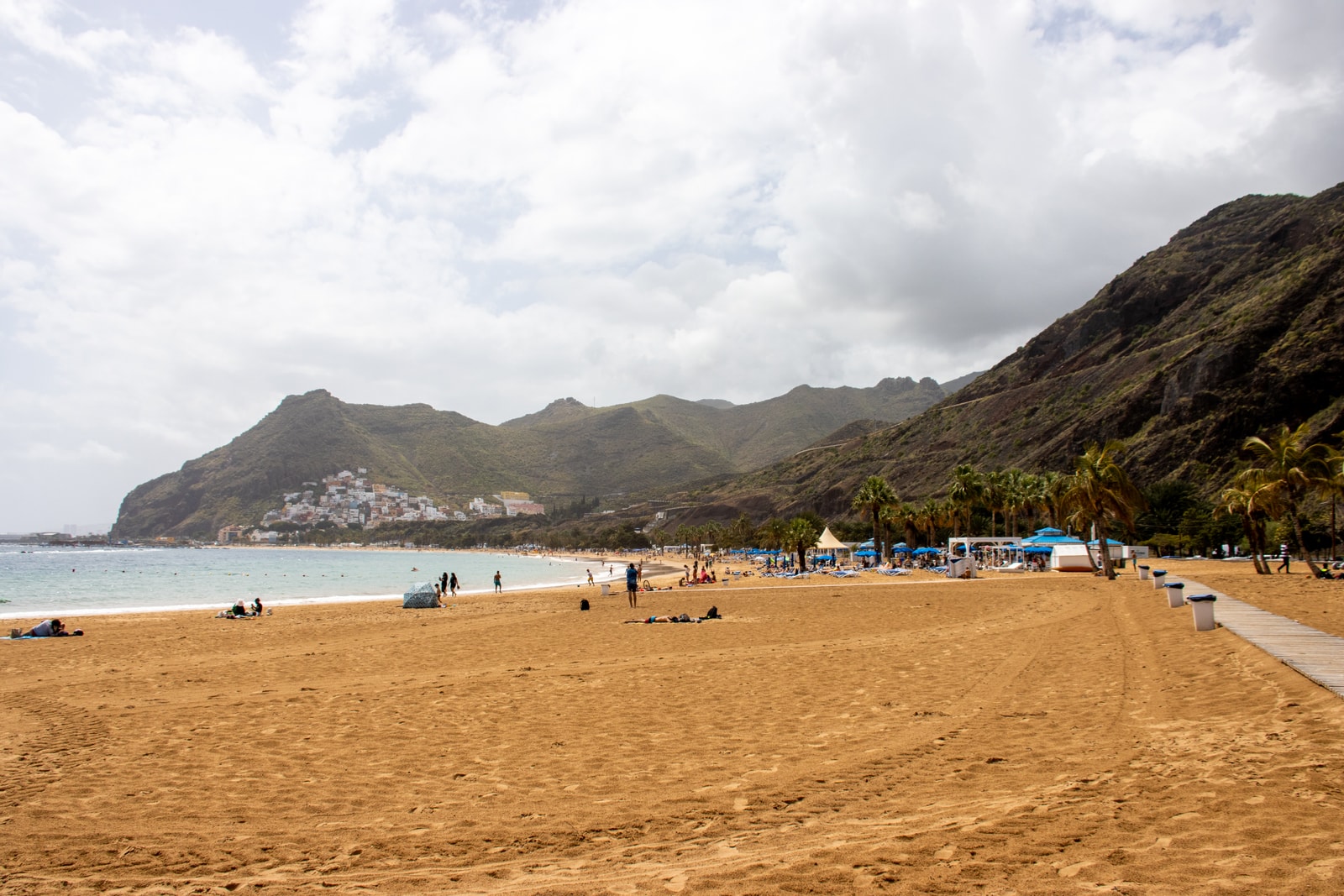 A ground-level view of Playa de Las Teresitas, showing its wide expanse of pale sand, a line of palm trees, and the rugged mountains behind it.