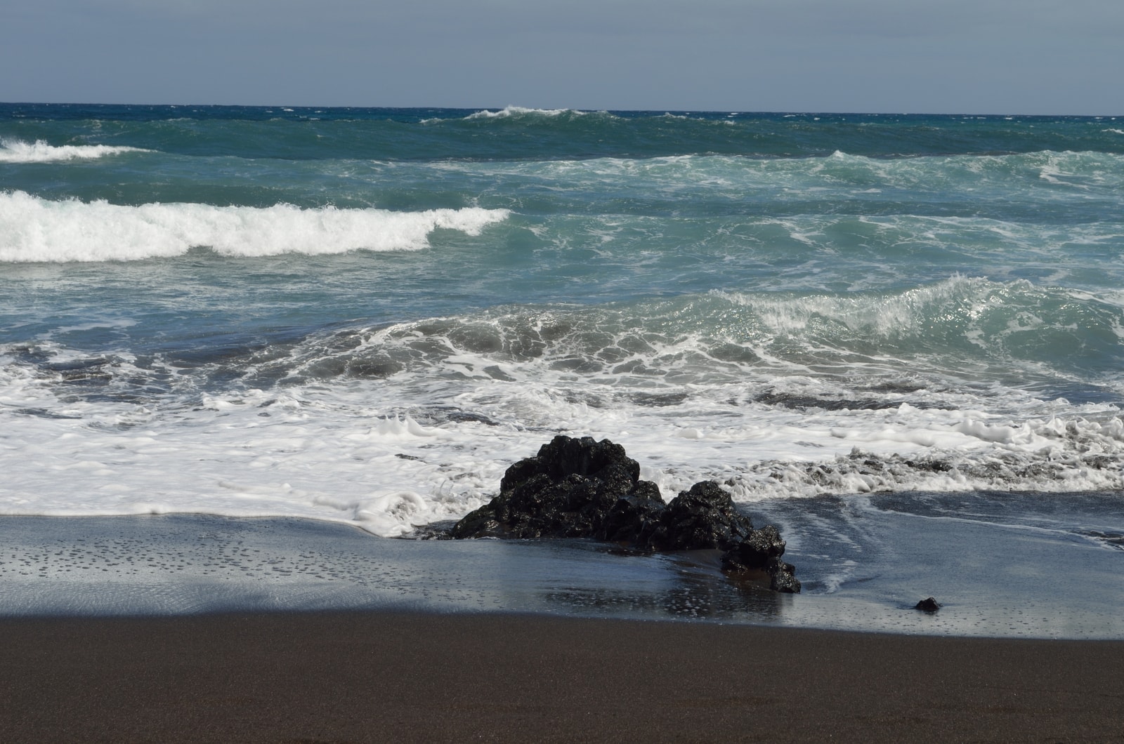 Foamy turquoise waves wash over a dark, textured volcanic rock on a beach.