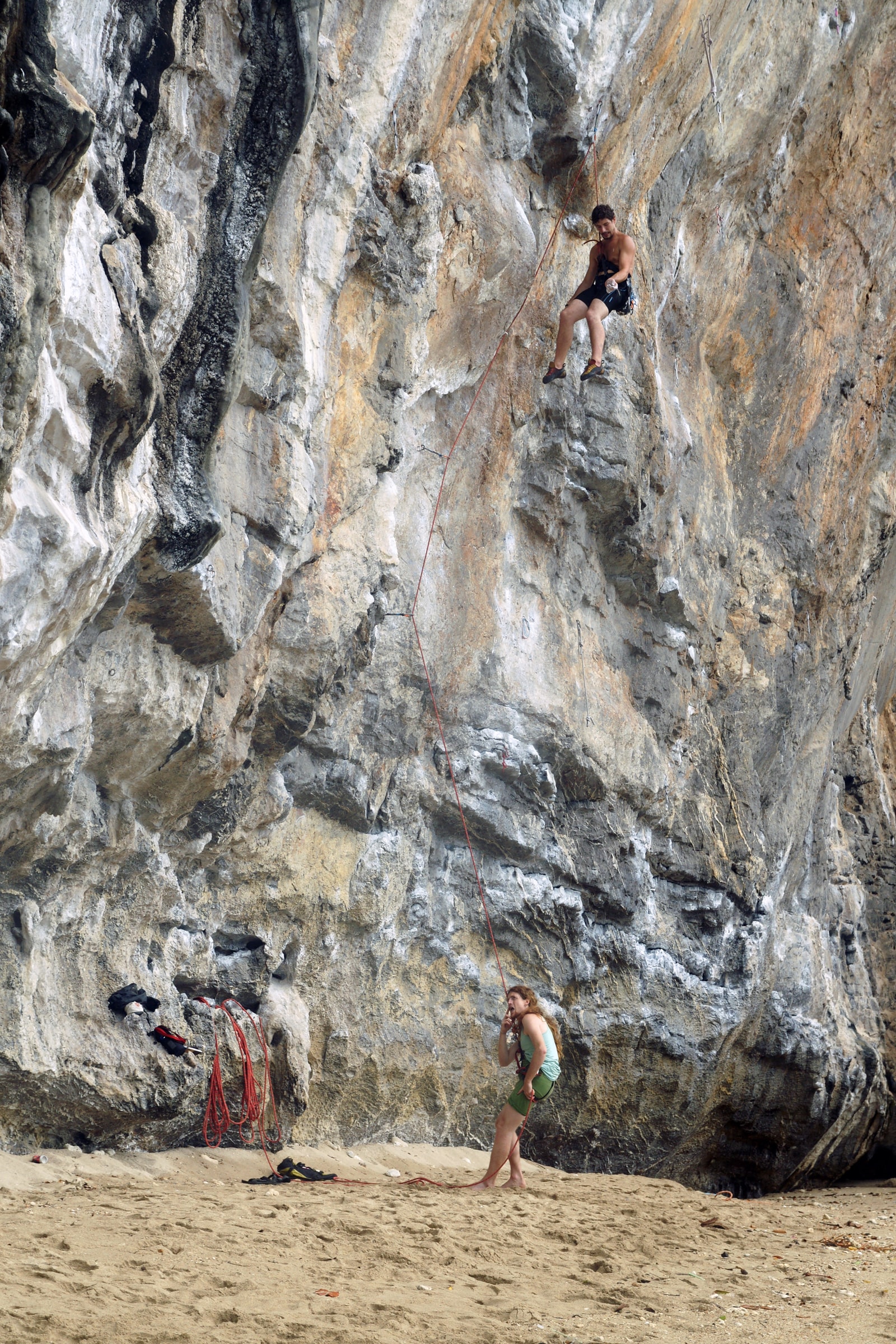 A rock climber scales a massive, textured limestone wall at Tonsai Beach, with their belayer visible at the bottom, showcasing the scale of the climb.
