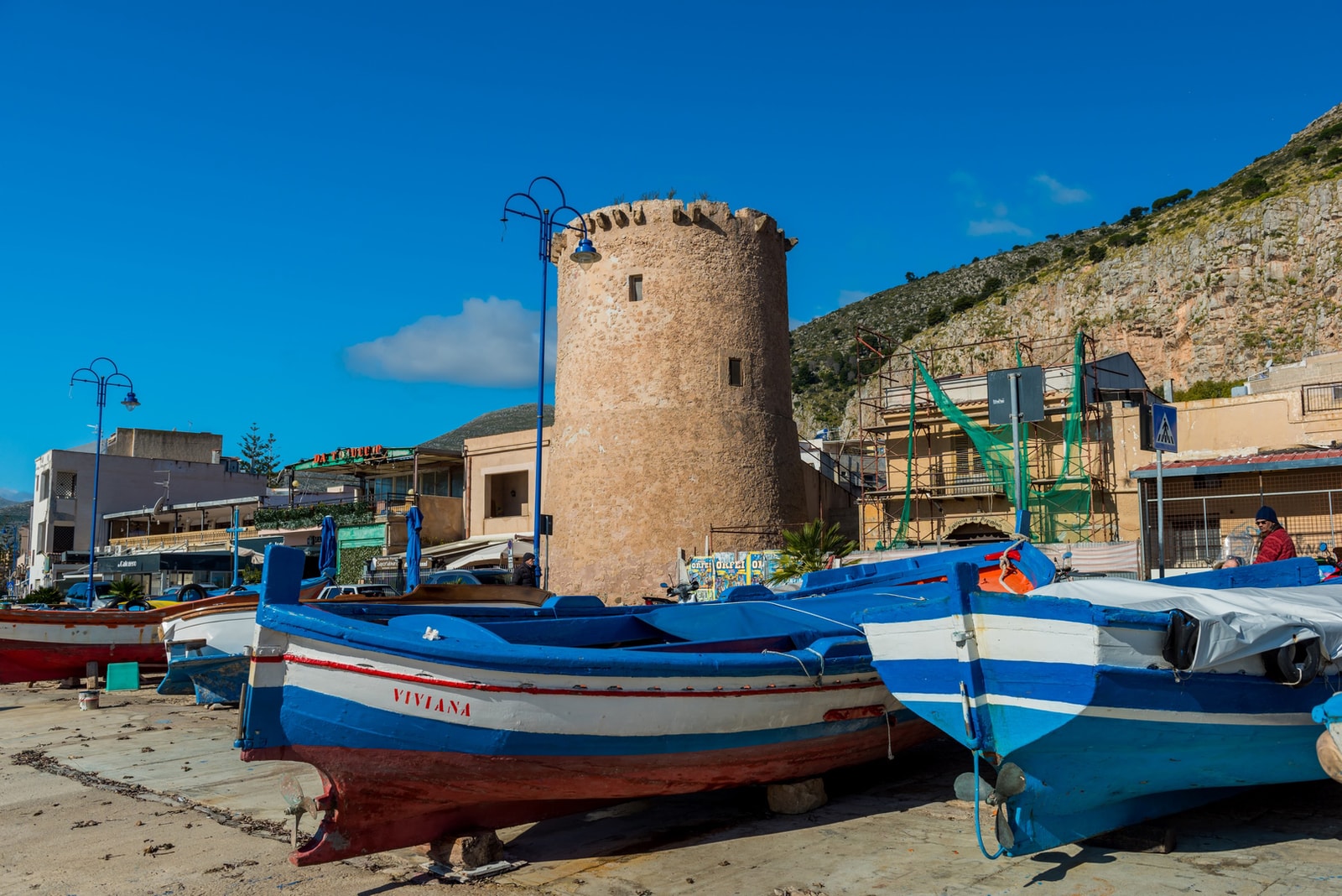 The historic stone watchtower, Torre di Mondello, stands at the edge of the harbor with traditional boats resting on the shore in front of it.