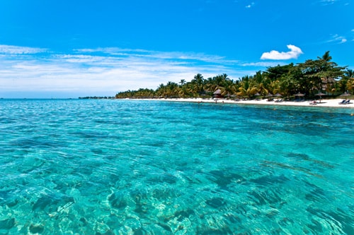 A beach scene at Trou aux Biches shows calm, clear turquoise water lapping against the white sand, with palm trees visible along the shoreline.