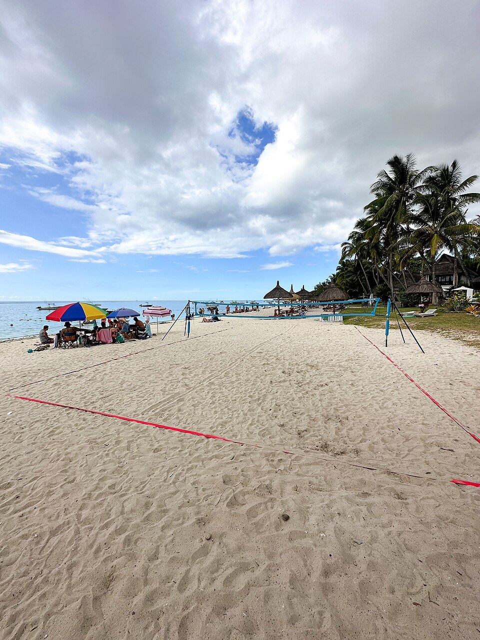 A beach volleyball net is set up on the sand at Trou aux Biches, with the calm blue lagoon and palm trees in the background.