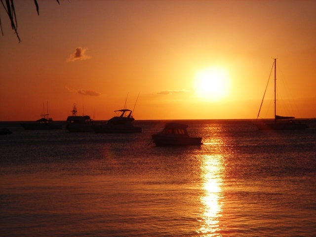 A vibrant orange and purple sunset over the ocean at Trou aux Biches, with the silhouettes of boats on the water.