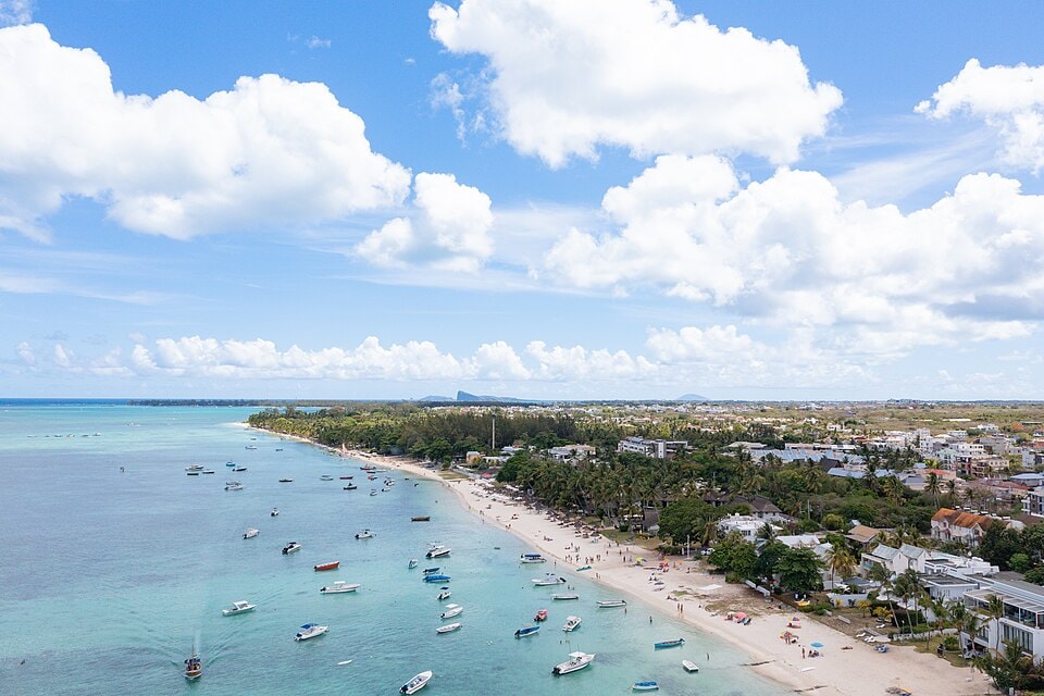 An aerial shot captures several yachts and boats anchored in the calm, vibrant blue lagoon of Trou aux Biches, with the beach and resorts in the background.