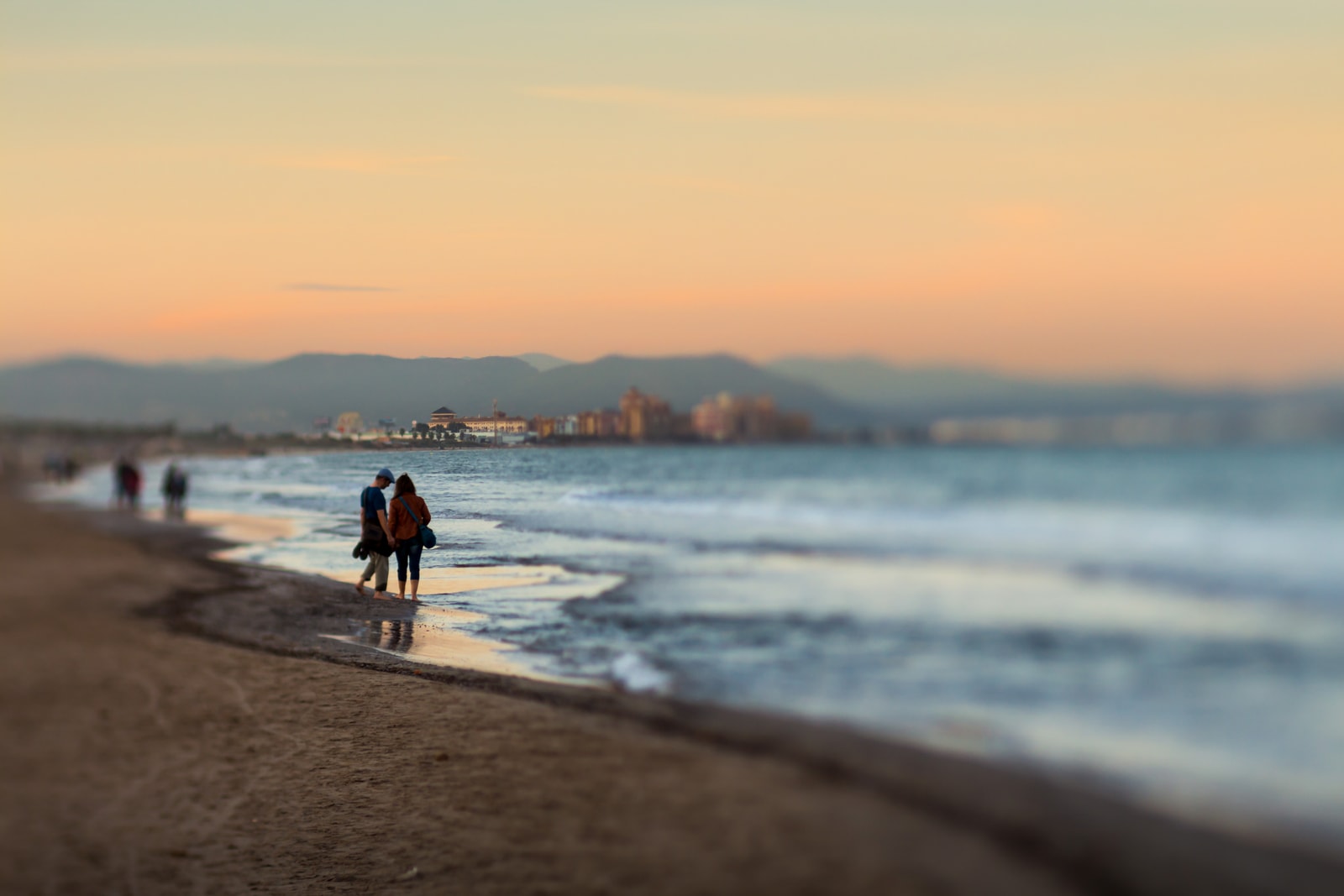 An artistic, soft-focus photo of a couple walking hand-in-hand along the shoreline at sunset.