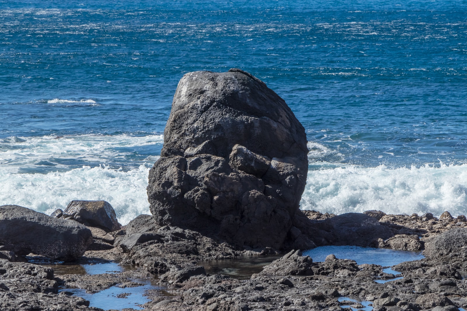 A dark volcanic boulder sits on a rocky shore with tide pools and surf in the background at Valle Gran Rey.